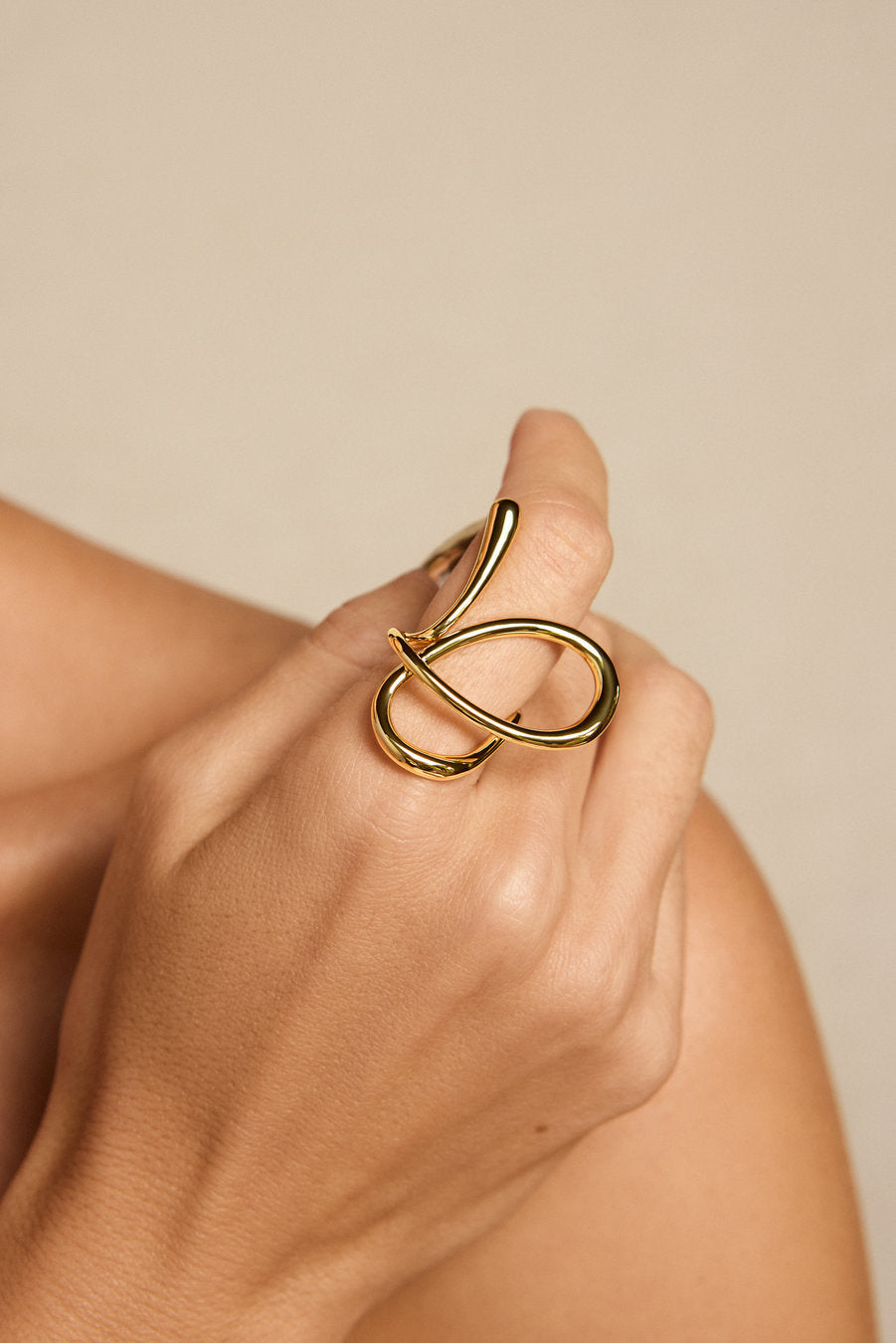 A close-up of a hand wearing a polished gold infinity-style ring, set against a soft beige background.
