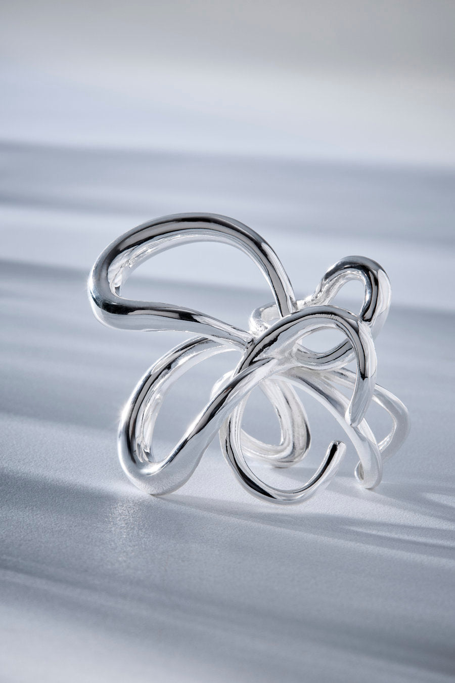 A polished silver abstract ring with flowing, intertwined loops, photographed on a soft light gray surface with a blurred studio background.