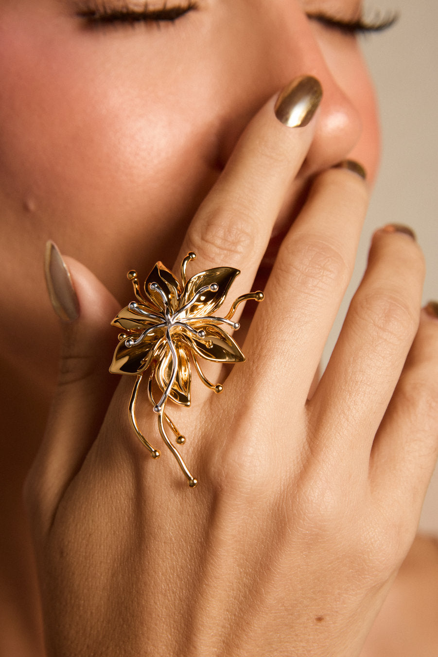 A close-up lifestyle shot of a woman's hand wearing a large, sculptural gold flower ring with silver accents on the center, set against her face.