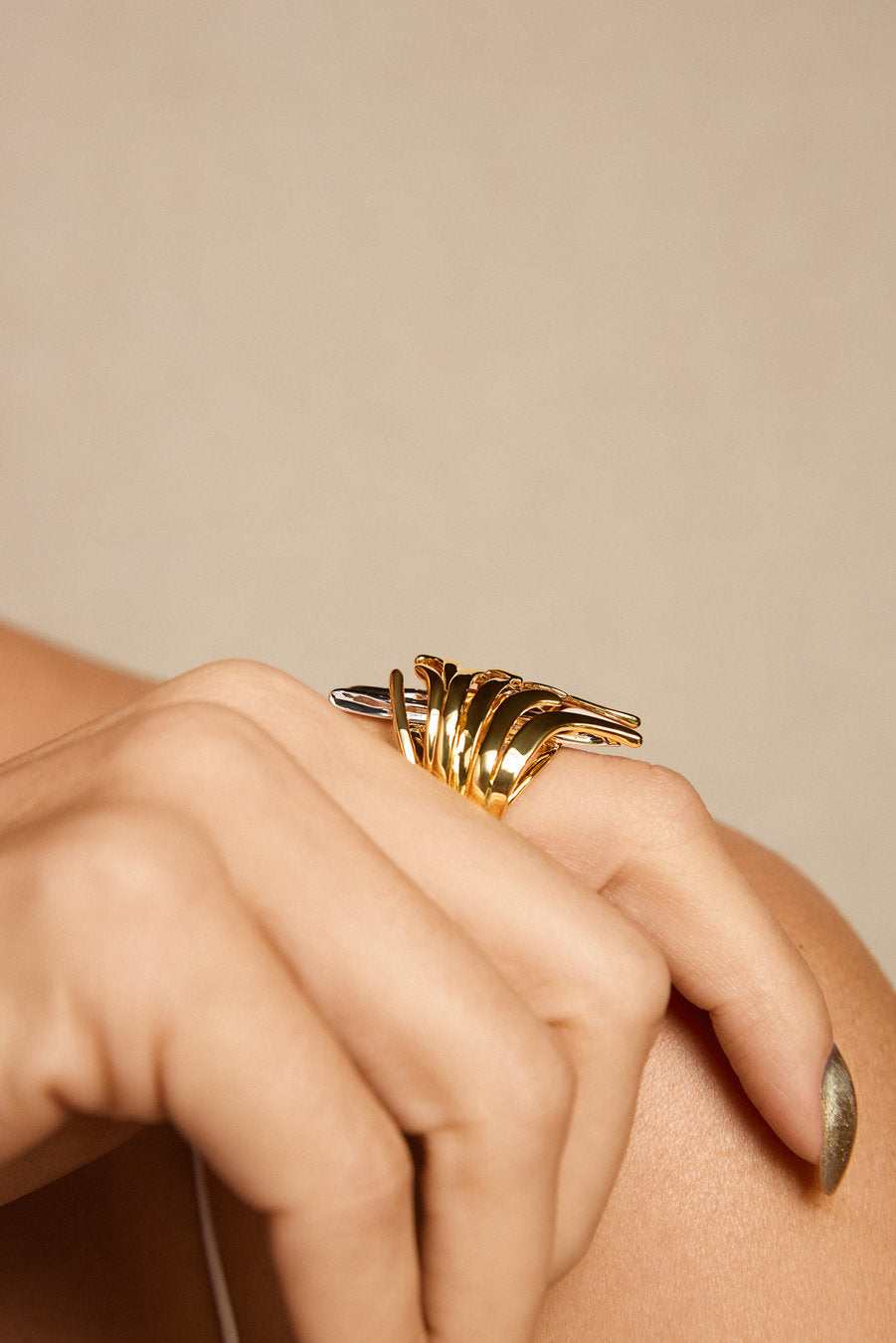 A close-up of a hand wearing a chunky, multi-strand gold ring with a textured, organic twisted design, set against a soft beige background.