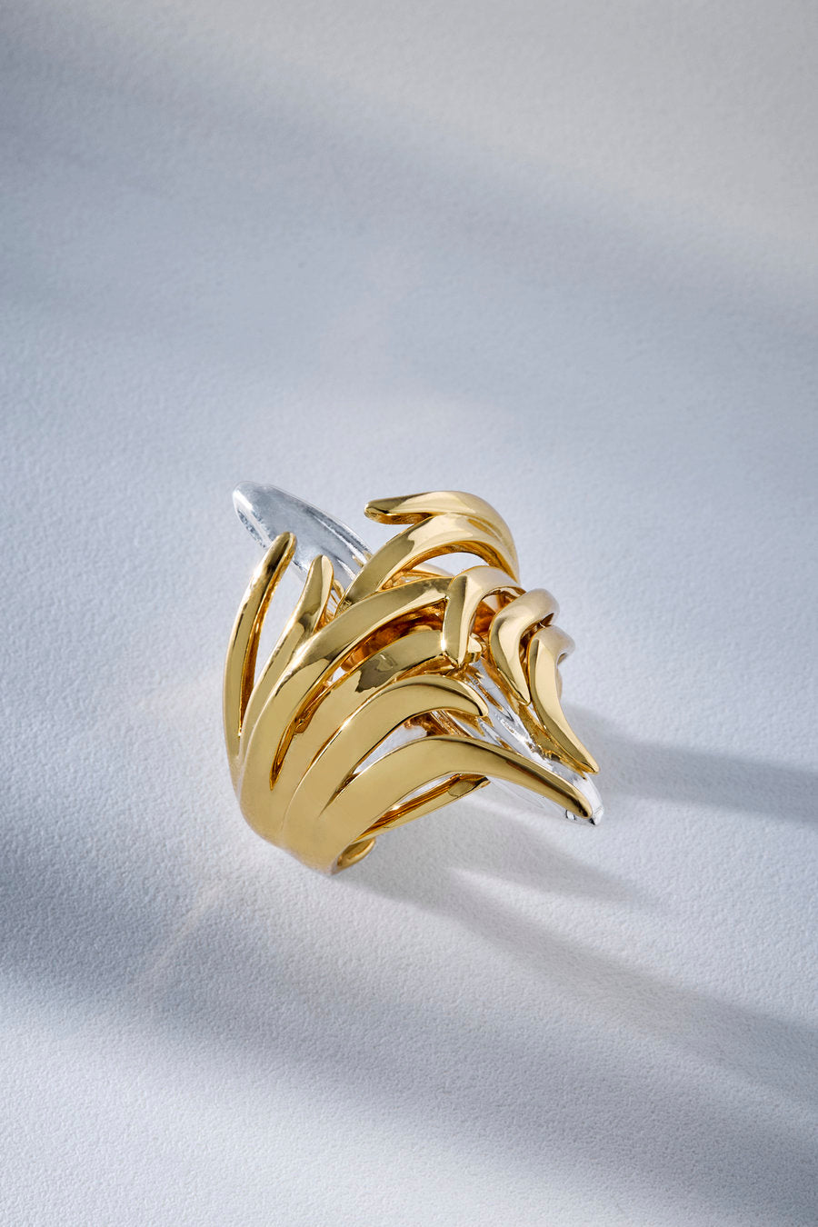 A two-tone gold and silver abstract wave ring with a polished finish, resting on a textured white surface with soft shadows.