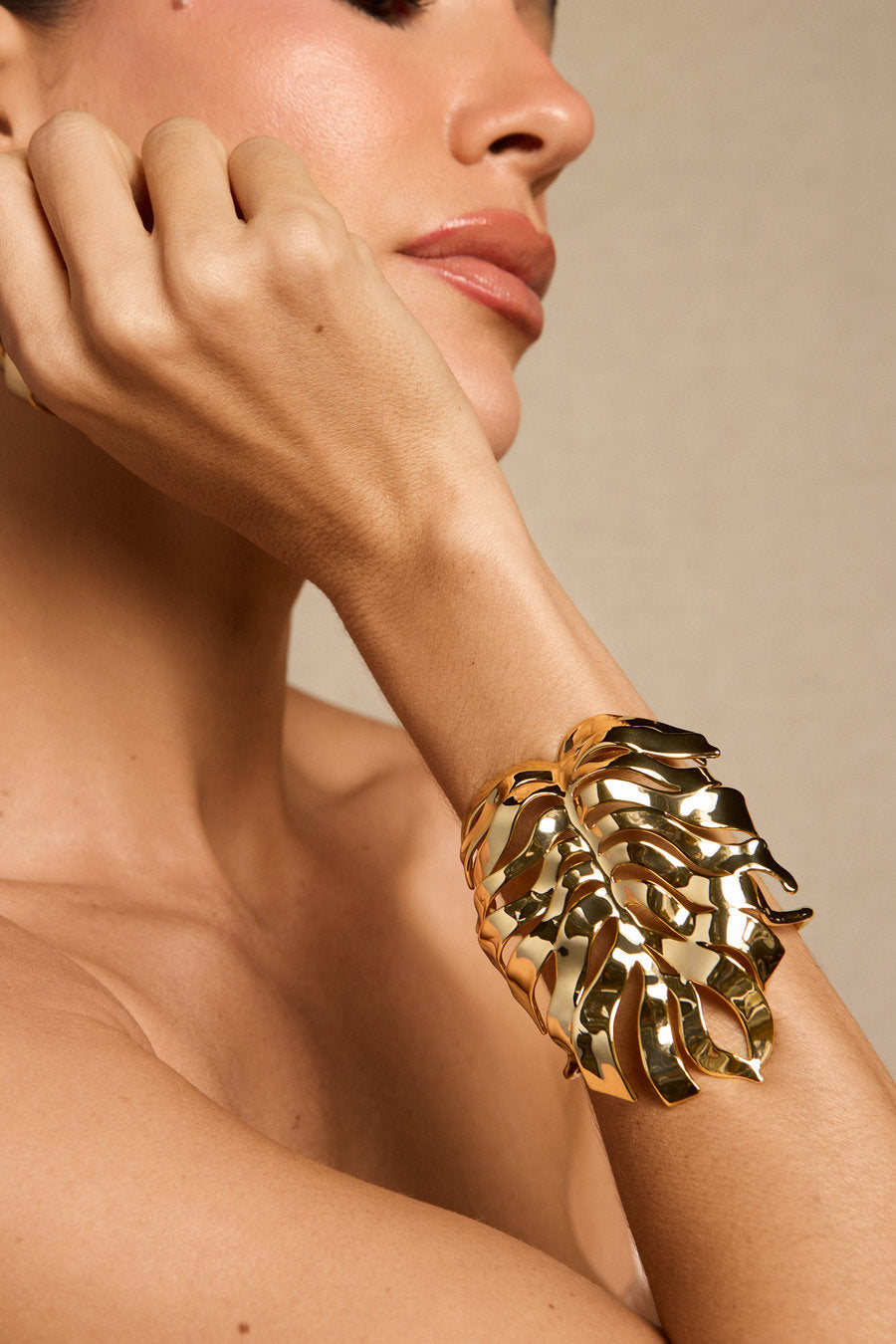 A close-up lifestyle shot of a model wearing a chunky, sculptural gold leaf cuff bracelet against a neutral beige background.