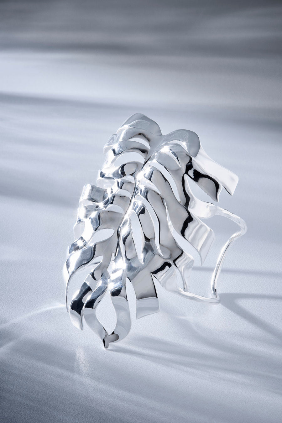 A sculptural, highly reflective silver ring with an organic, flowing, leaf-like or flame-like layered design, photographed on a textured light gray surface.