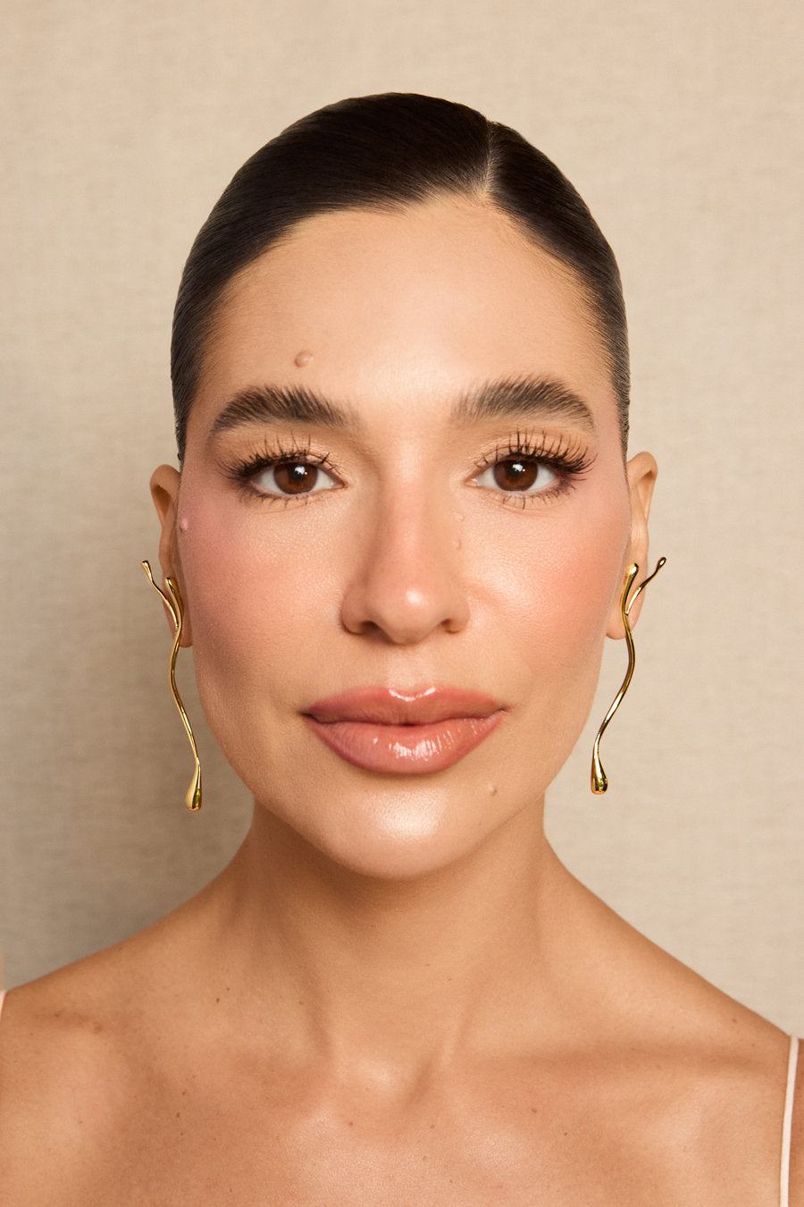 A close-up portrait of a woman with her hair pulled back, wearing large, sculptural gold hoop earrings with a wavy, fluid design. The background is a soft, textured beige wall.