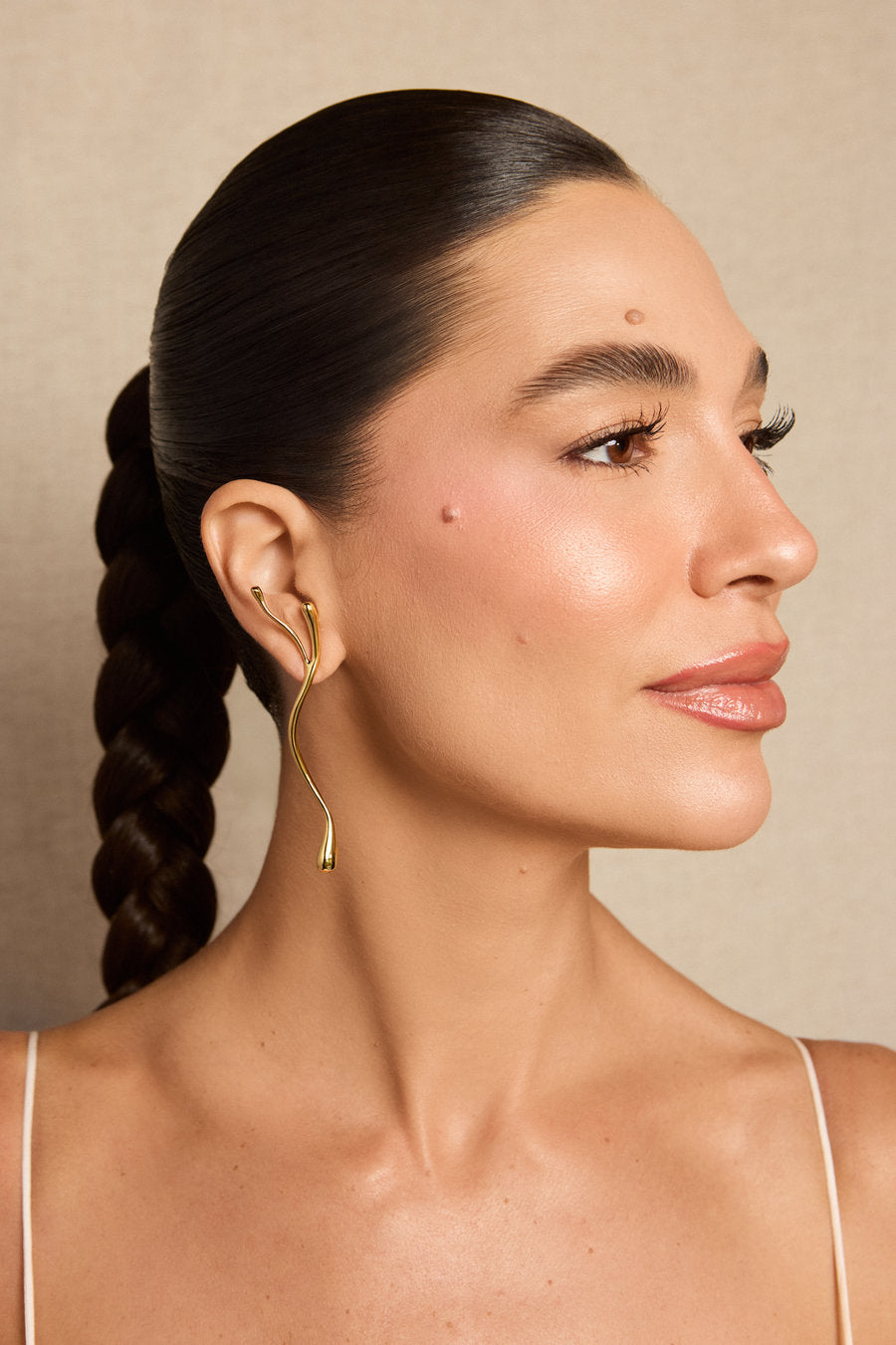 A woman in profile wearing a delicate gold wavy hoop earring, shown against a neutral beige background.