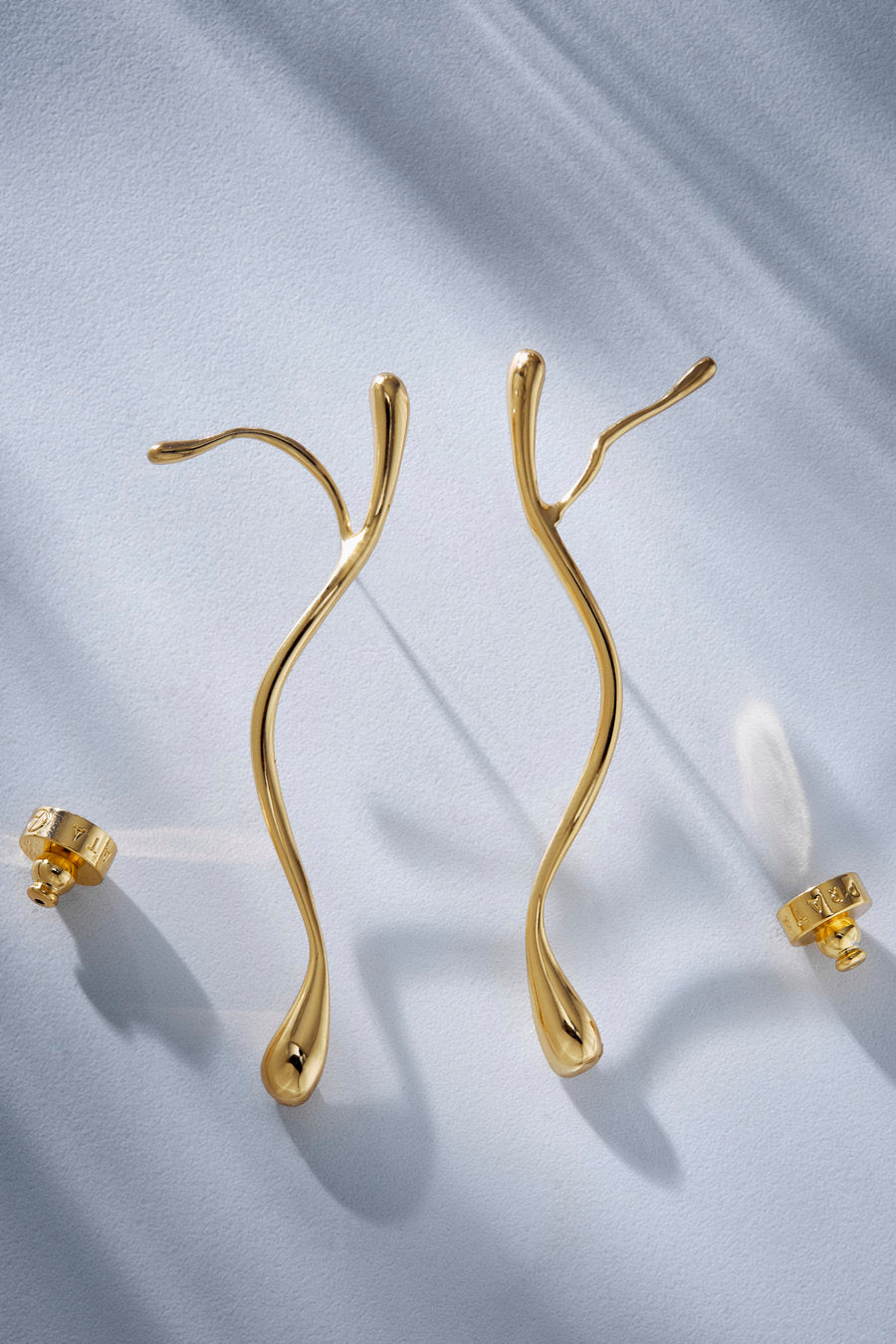 A pair of gold abstract wavy drop earrings with post backings, displayed on a light blue textured fabric background with soft shadows.