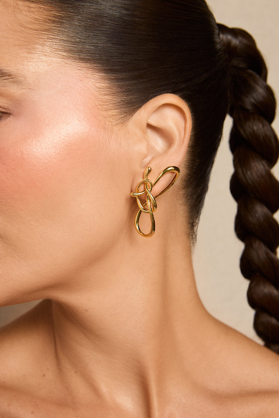 A close-up side profile of a woman wearing a gold abstract loop earring, with her dark hair styled in a braid against a neutral beige background.