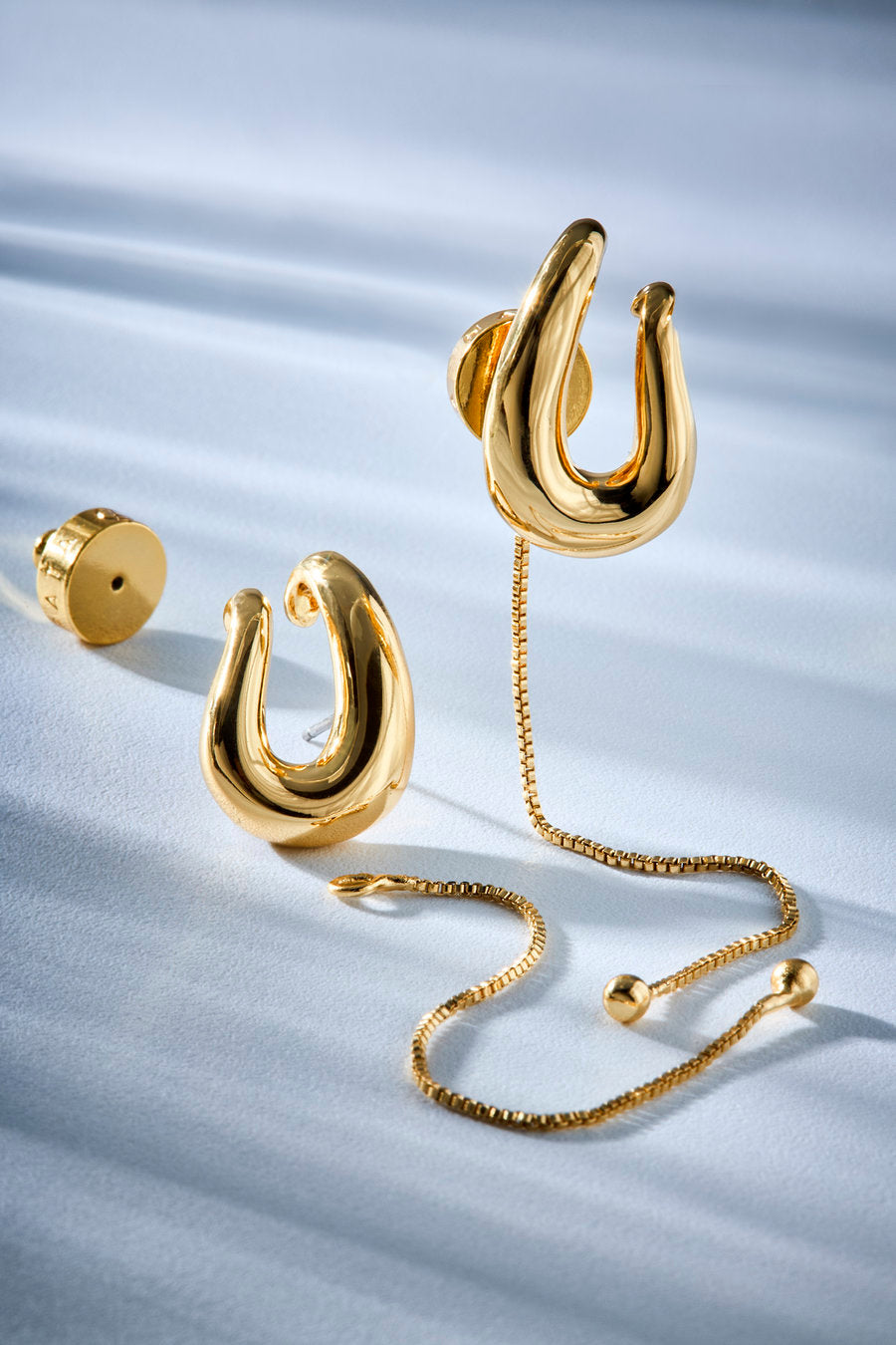 Gold hoop-style earrings with a matching chain, shown on a light blue fabric background. One earring is displayed upright, another lies on its side, and the chain drapes across the surface.