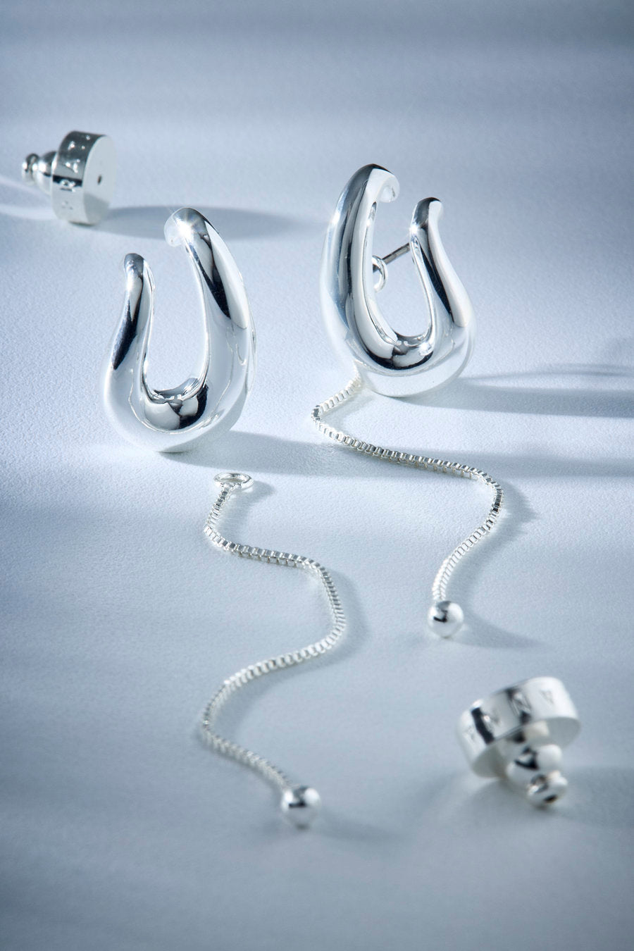 A pair of polished silver earrings with abstract curved hooks and dangling chain links, displayed on a clean light gray surface.