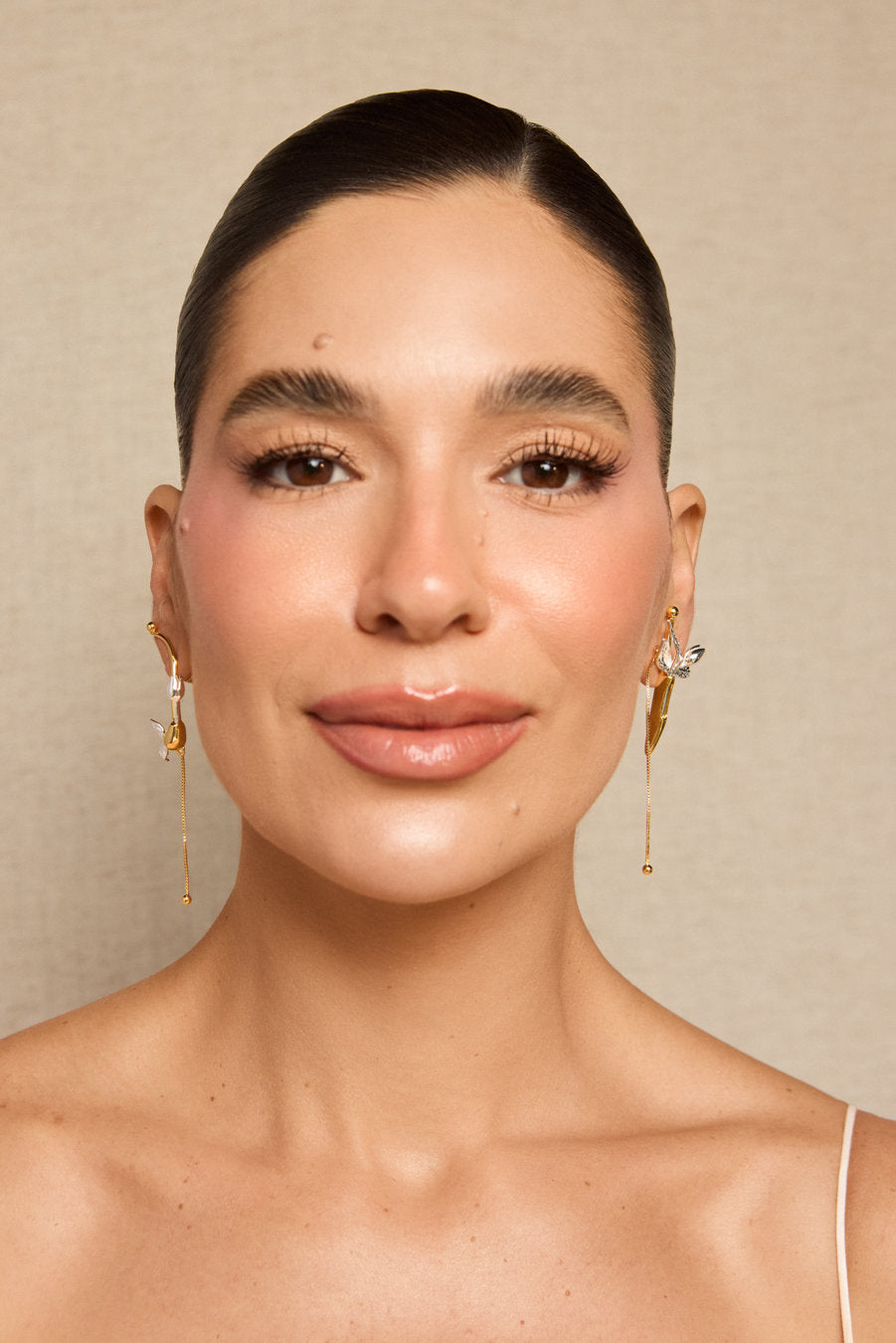 A close-up portrait of a woman wearing elegant gold dangle earrings with clear stones, set against a neutral beige textured background.