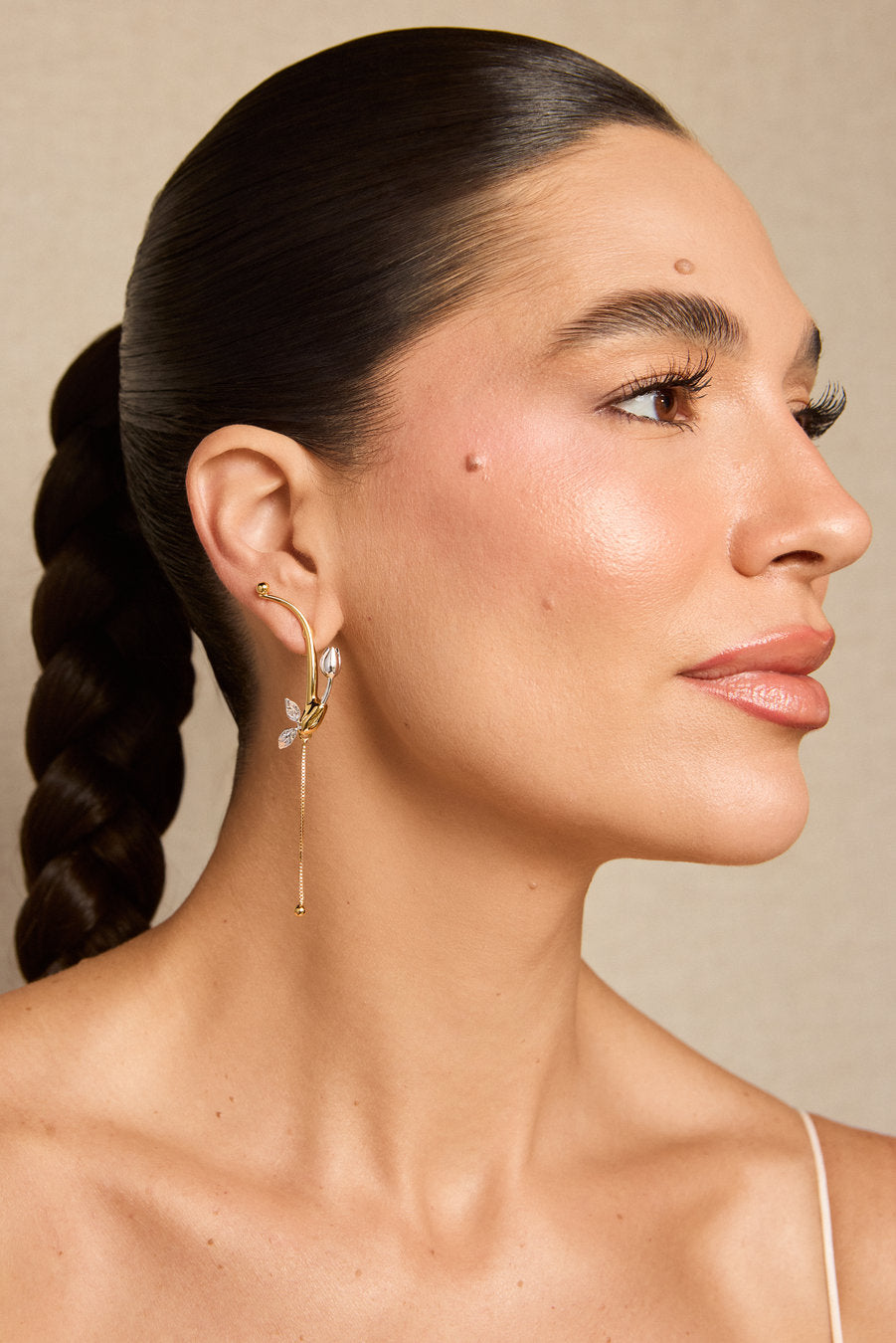 A close-up side profile of a woman with dark hair in a braid, wearing a delicate gold hoop earring with a dangling leaf-shaped charm, against a warm beige background.