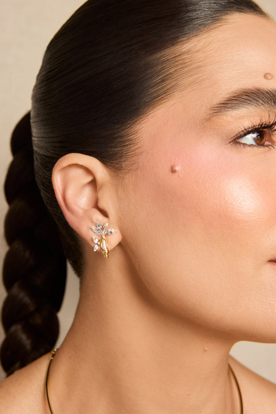 A close-up of a model's ear wearing a gold and crystal flower-shaped stud earring, with her hair in a dark braid against a neutral beige background.