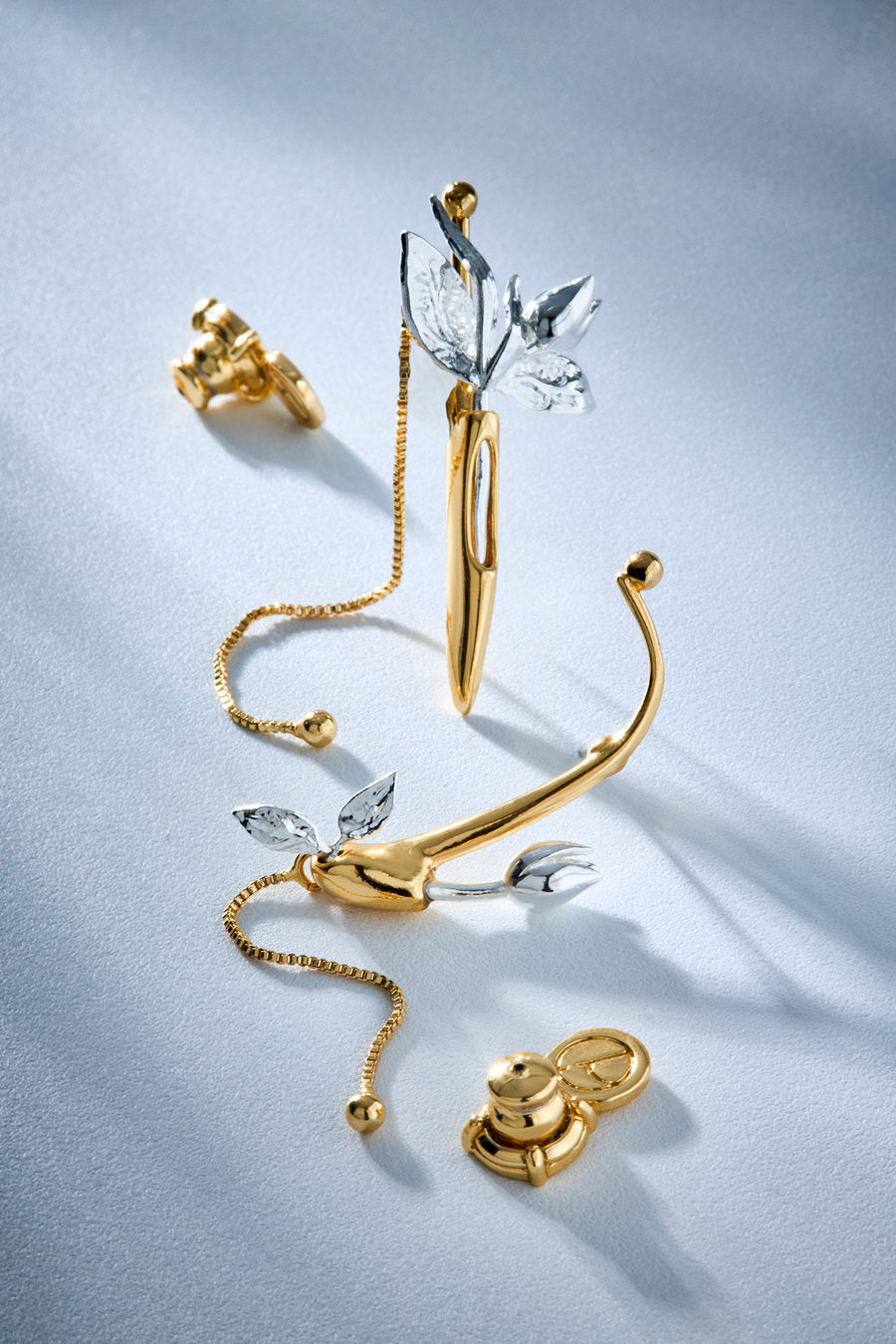 A pair of gold and silver leaf-shaped hoop earrings with a delicate gold chain, displayed on a light gray textured background.