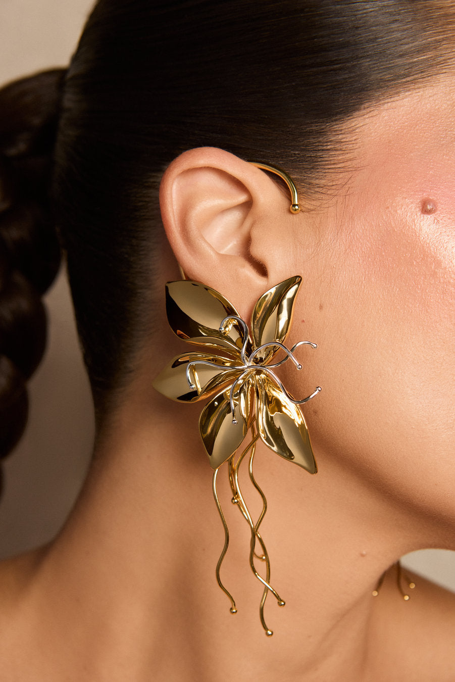 A close-up of a model's ear wearing a large, sculptural gold flower-shaped earring with dangling wavy gold wires, alongside a smaller gold hoop earring.
