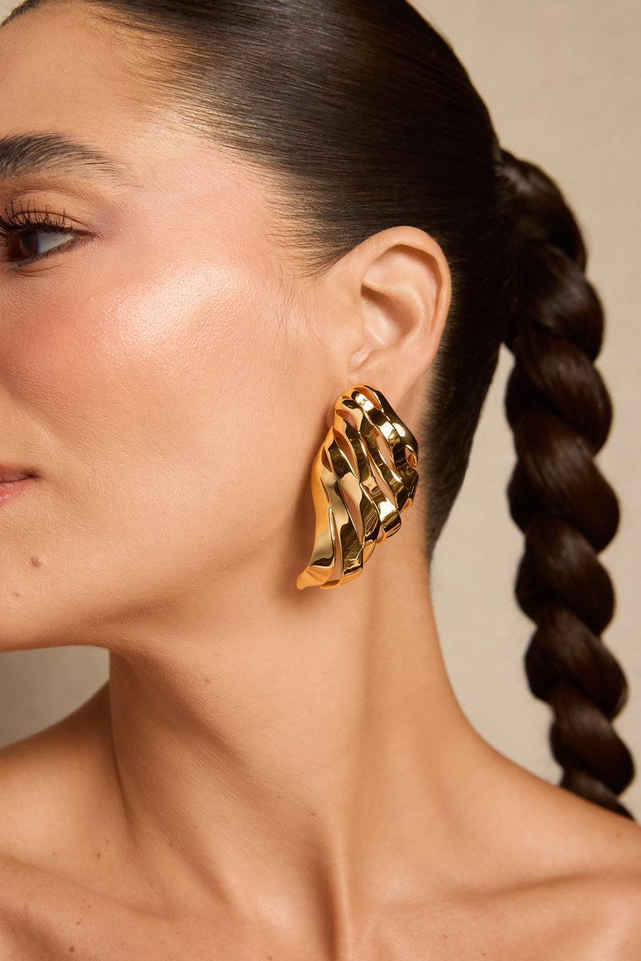 A close-up of a model wearing a large, chunky gold earring with a twisted, layered design. The model has dark brown hair styled in a side braid against a neutral beige background.