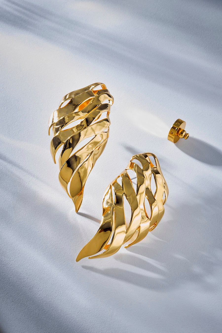 A pair of gold leaf-shaped earrings with a polished finish, displayed on a white textured fabric background with soft shadows.
