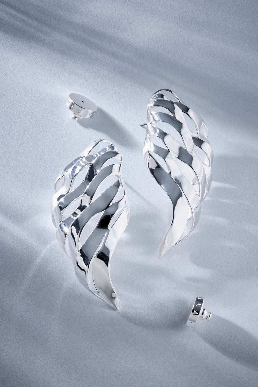 A pair of sculptural, polished silver earrings with a layered, wavy design, displayed on a textured light gray fabric background with their post backs visible.
