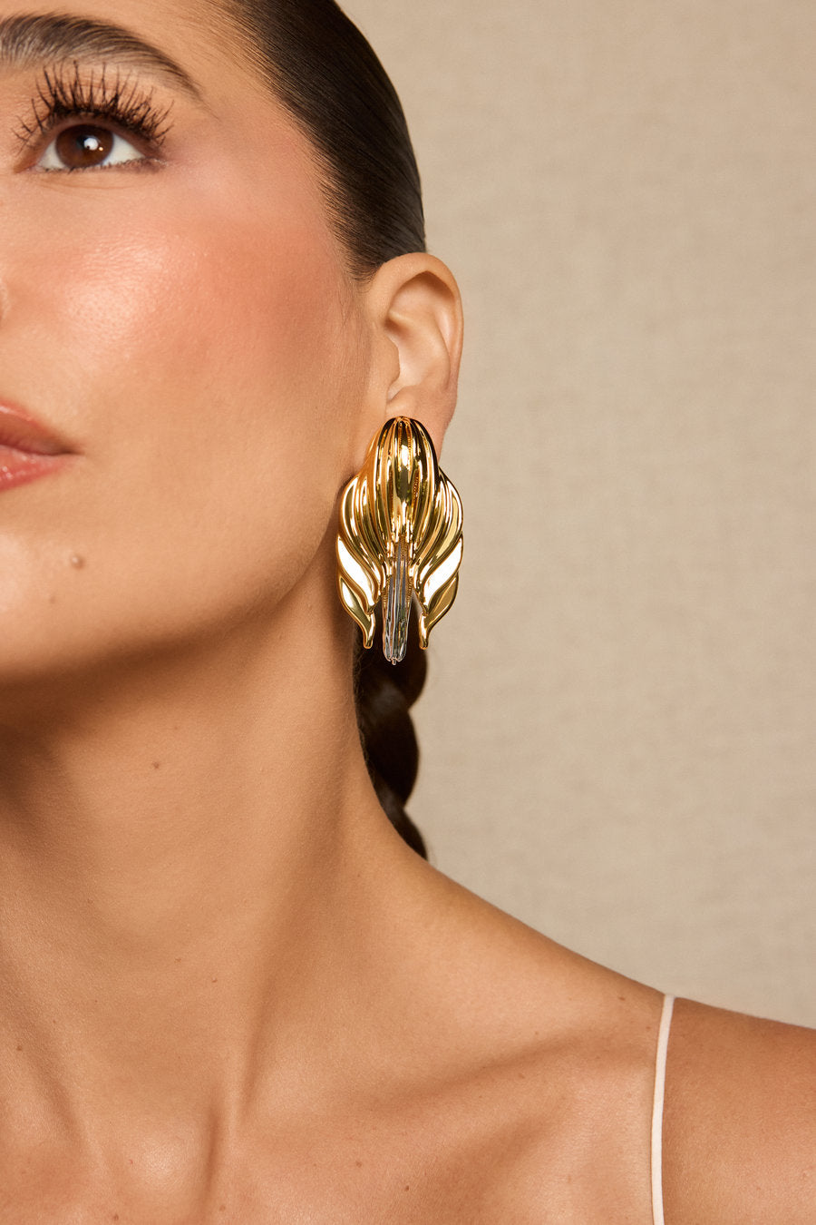 A close-up of a woman wearing a large, sculptural gold earring with layered leaf-like petals and a central clear stone, set against a neutral beige background.