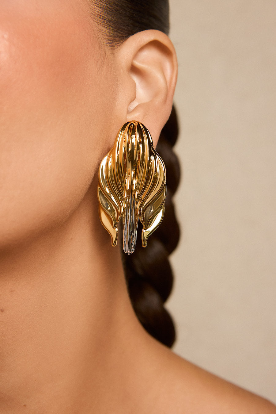 A close-up of a model's ear wearing a large, sculptural gold earring with layered leaf-like or petal-like shapes and a clear faceted center stone, against a warm beige background.
