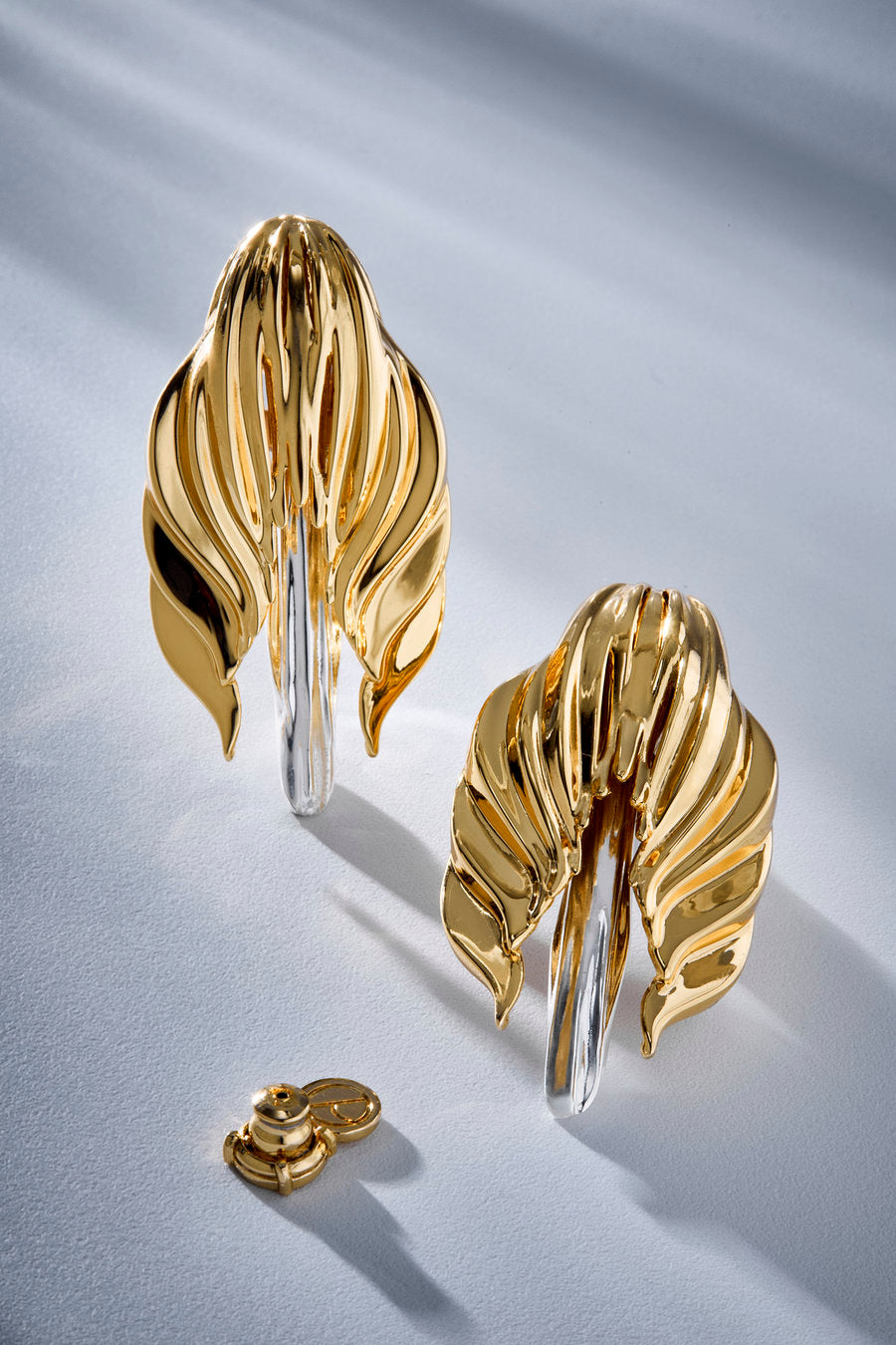 A pair of sculptural gold earrings with a leaf-like layered design and a silver-toned vertical post, shown on a light gray surface with soft shadows and a small detached gold clasp.