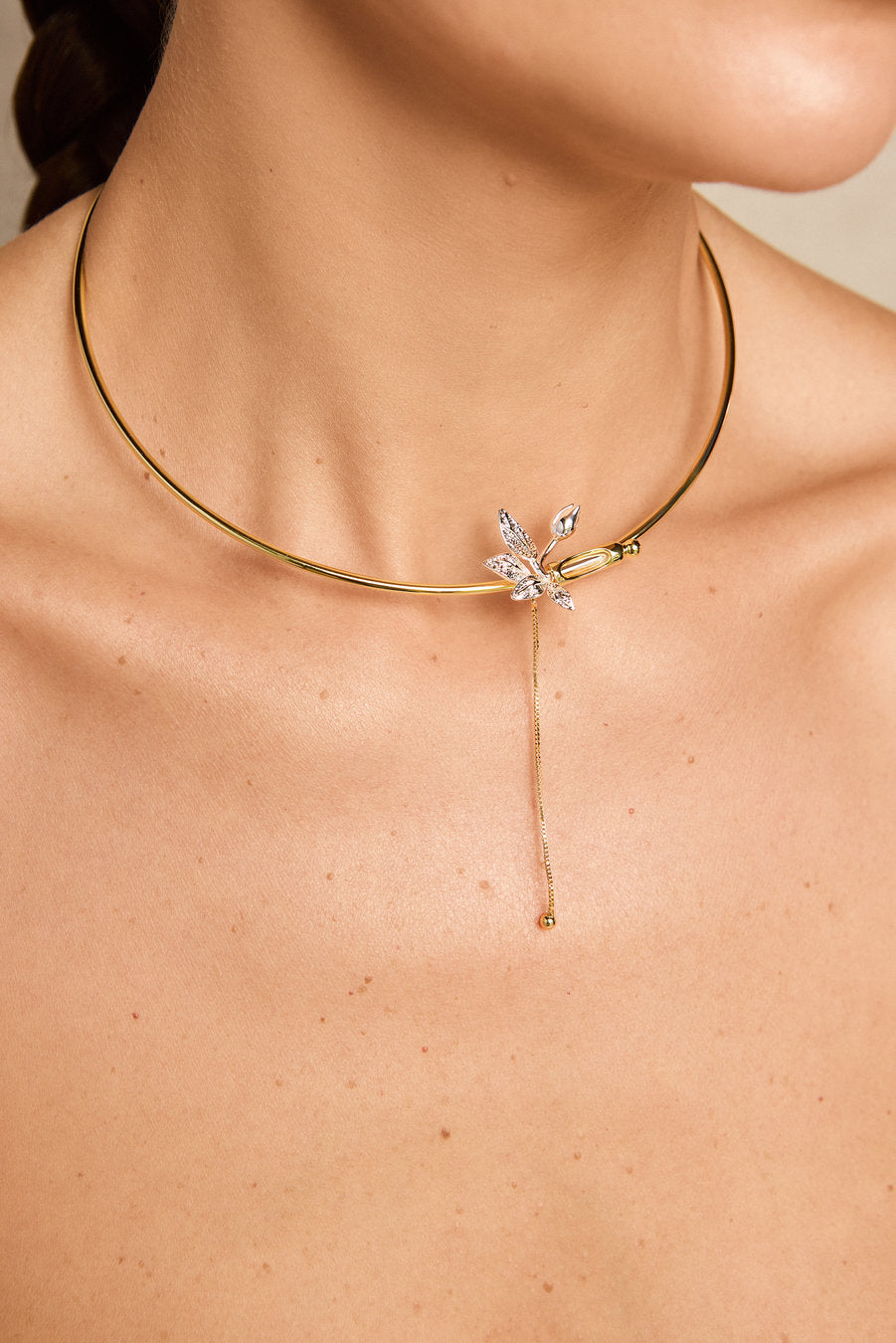 A close-up of a model's neck and upper chest wearing a delicate gold choker necklace with a small crystal-embellished flower and a long drop chain.