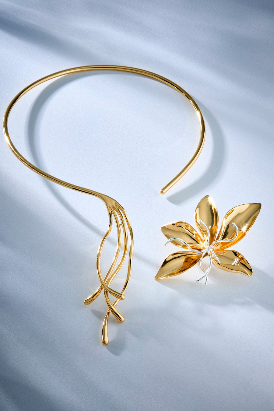 A gold-toned open cuff necklace with a leaf-like drape and a matching flower-shaped brooch, displayed on a light blue satin fabric background.