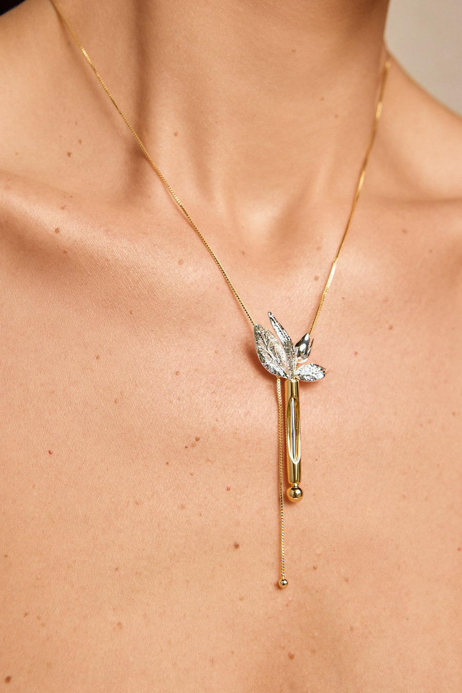 A close-up of a model's neck and chest wearing a delicate gold chain necklace with a leaf-shaped pendant featuring clear crystals and a vertical gold bar with a small bead at the bottom.