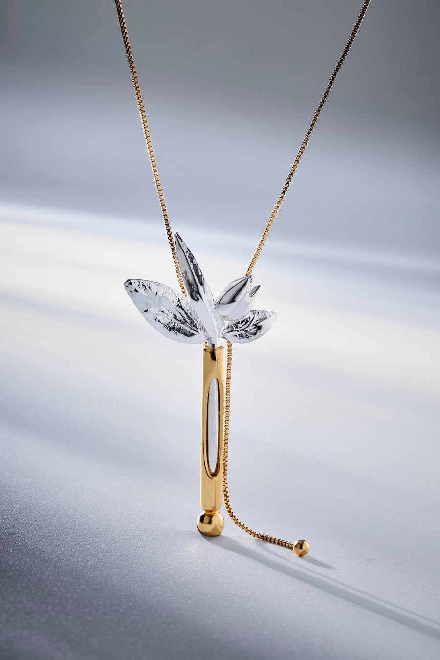 A gold and silver dragonfly pendant necklace on a delicate gold chain, displayed against a soft gray background with a subtle reflection.