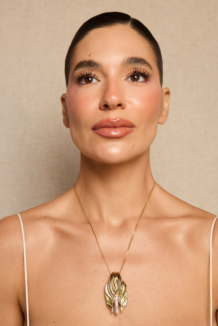 A close-up portrait of a woman wearing a delicate gold chain necklace with a textured, leaf-like gold pendant. She is shown from the chest up against a neutral beige background.