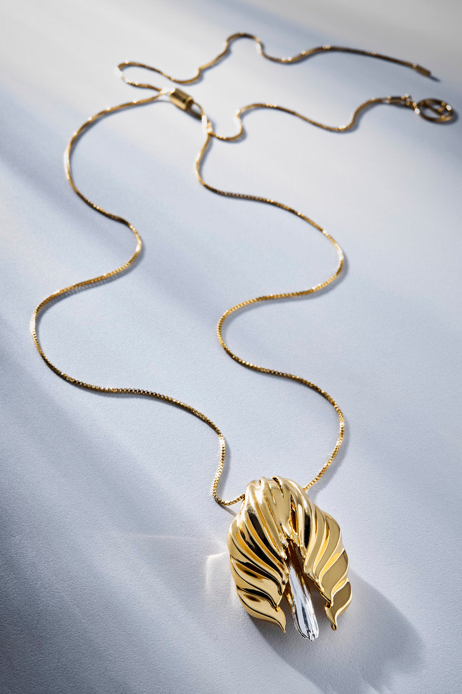 A gold pendant necklace with a sculptural, leaf-like form and a clear faceted crystal accent, displayed on a white textured fabric background.