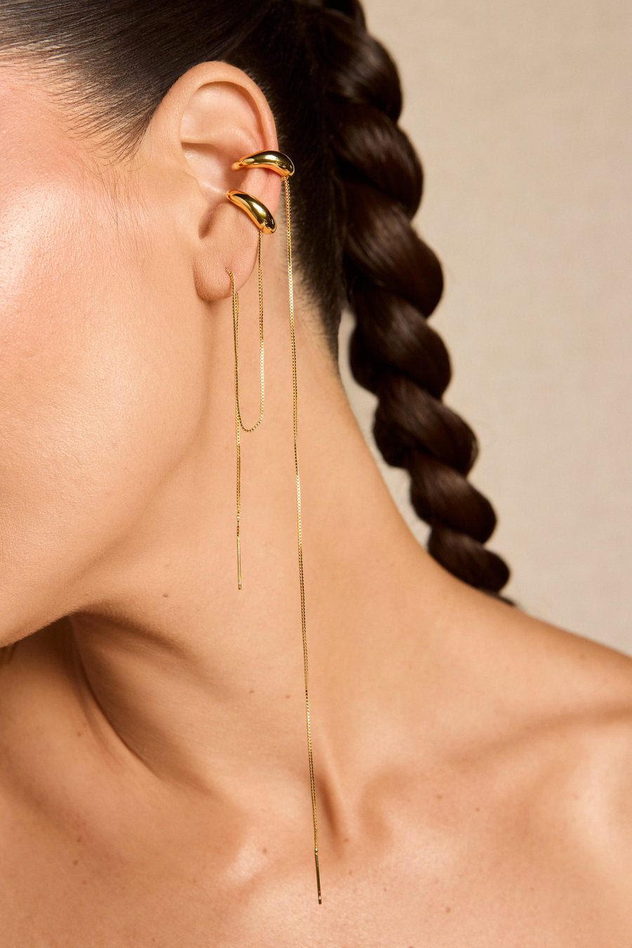 A close-up of a model's ear wearing a pair of gold ear cuffs with long dangling chain earrings, against a neutral beige background.