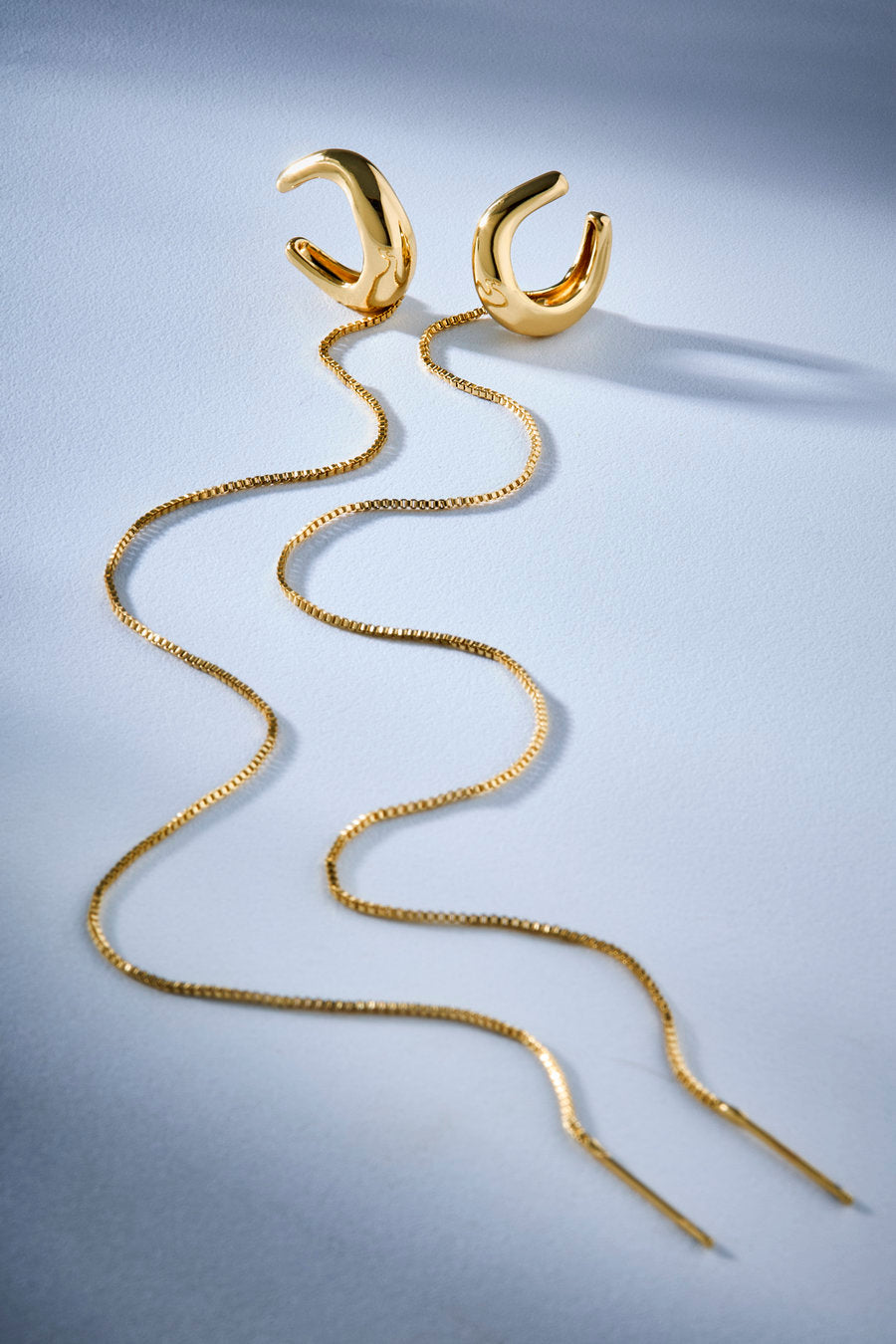 A pair of gold huggie hoop earrings with long, wavy snake-chain drop tails, displayed on a light blue-gray background.