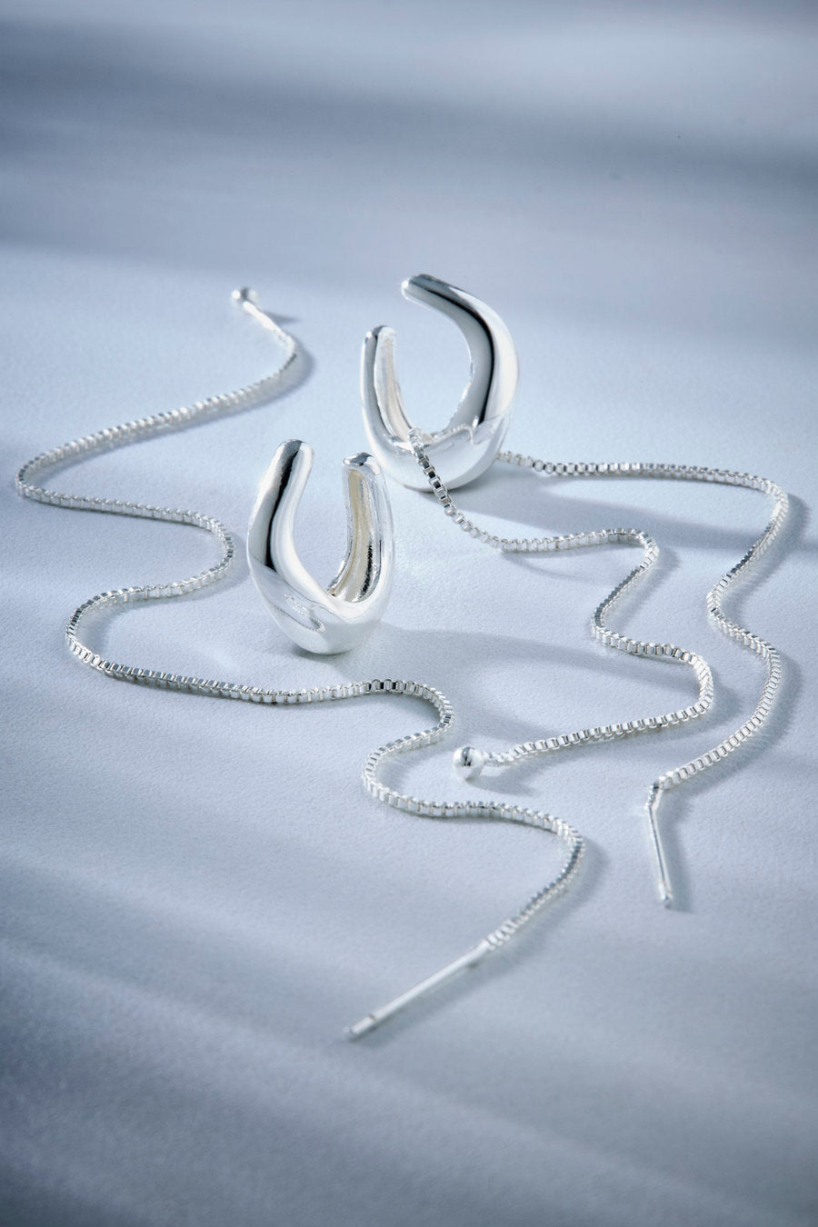 A pair of polished silver hoop-style earrings with matching silver chains, arranged on a light blue-gray satin fabric background.