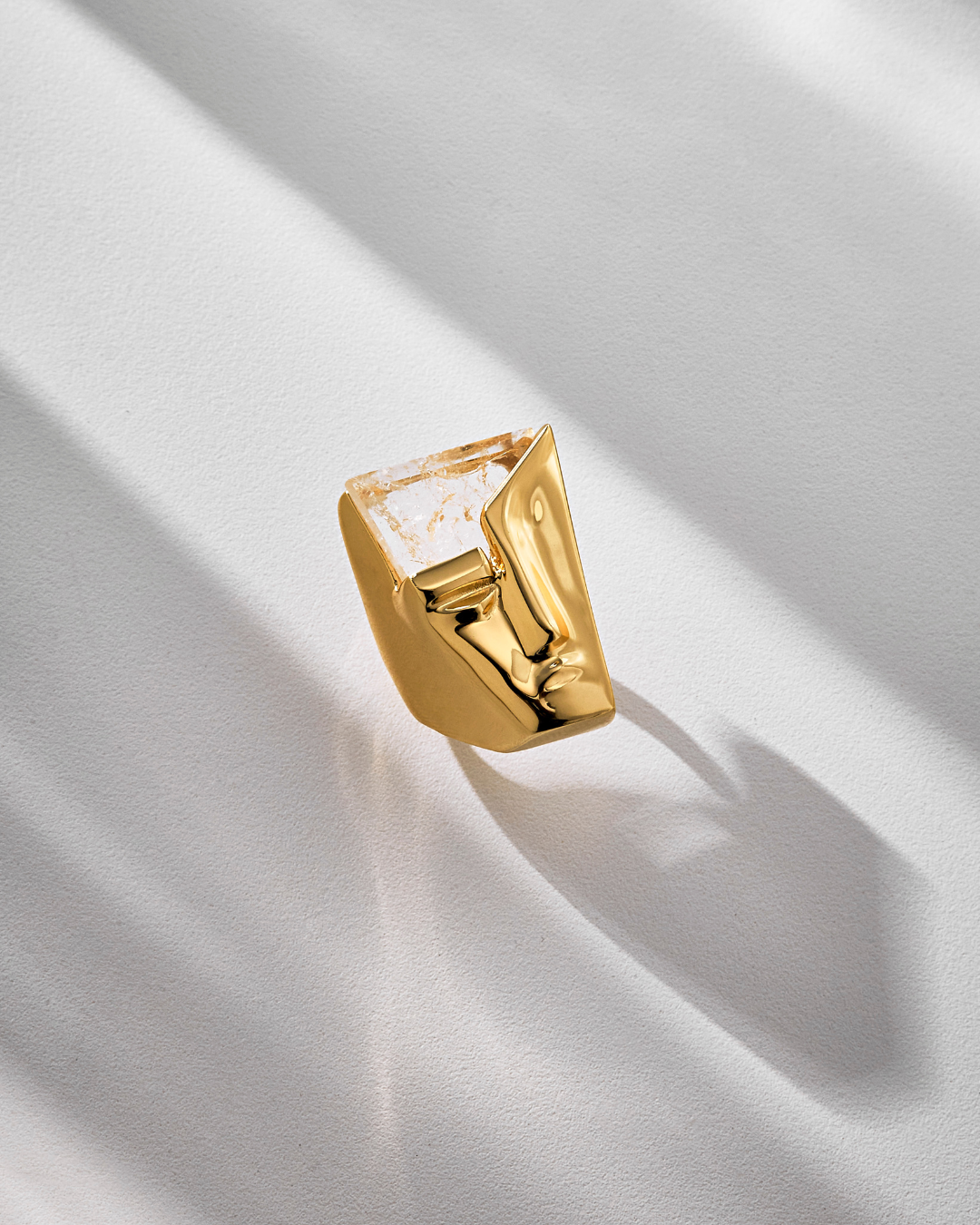 A gold-colored ring with a geometric design, featuring a quartz crystal, placed on a white surface with a shadow cast over it.
