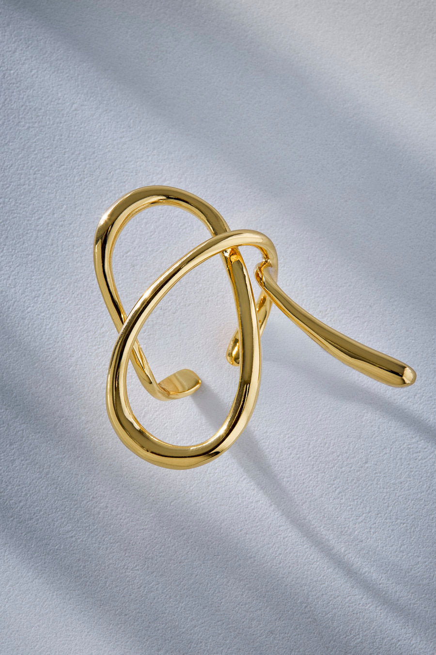 A polished gold abstract knot ring brooch with a smooth, reflective finish, displayed on a textured light gray fabric background.