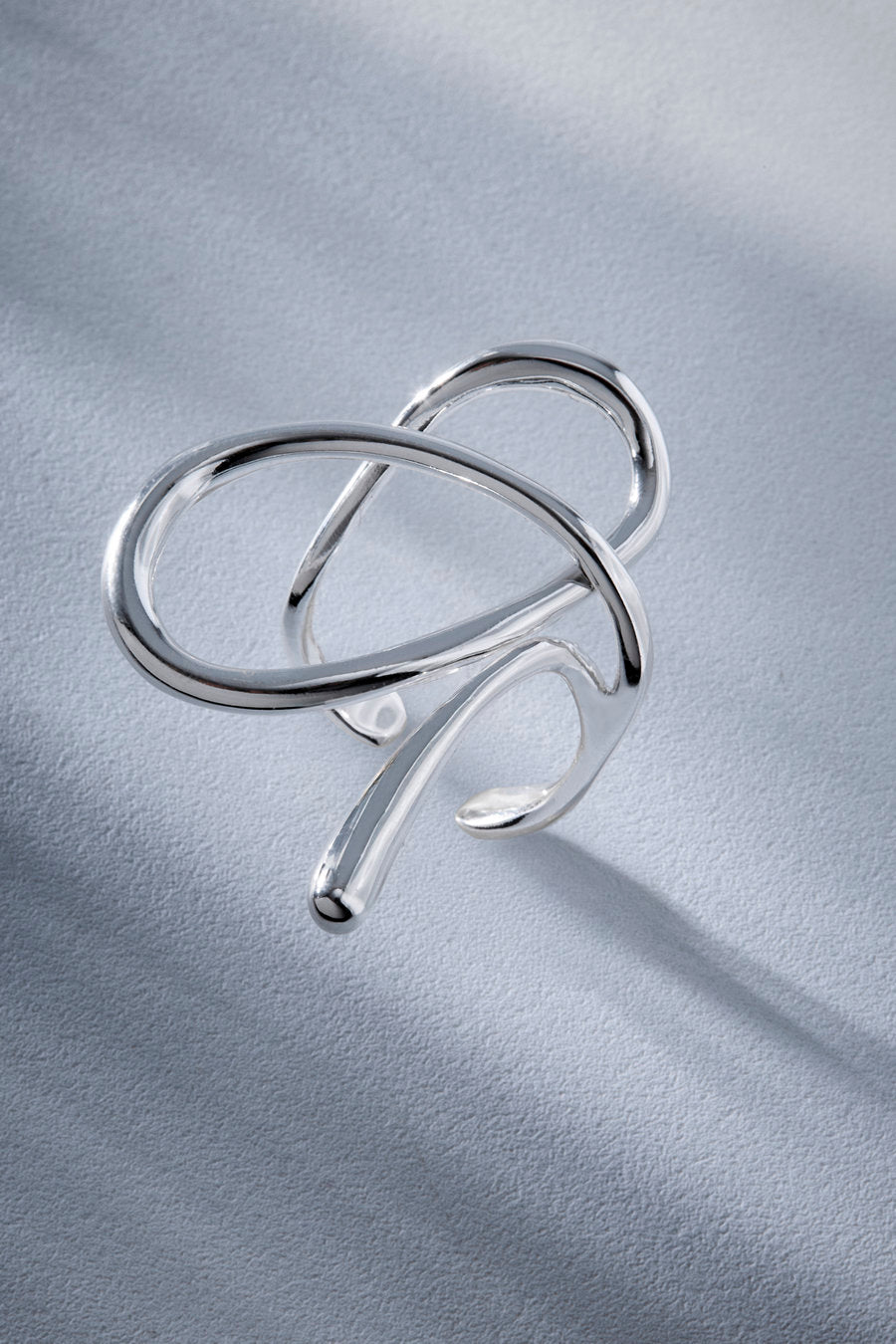 A polished silver-toned open cuff ring with an abstract, flowing looped design, displayed on a textured light gray surface with soft shadows.