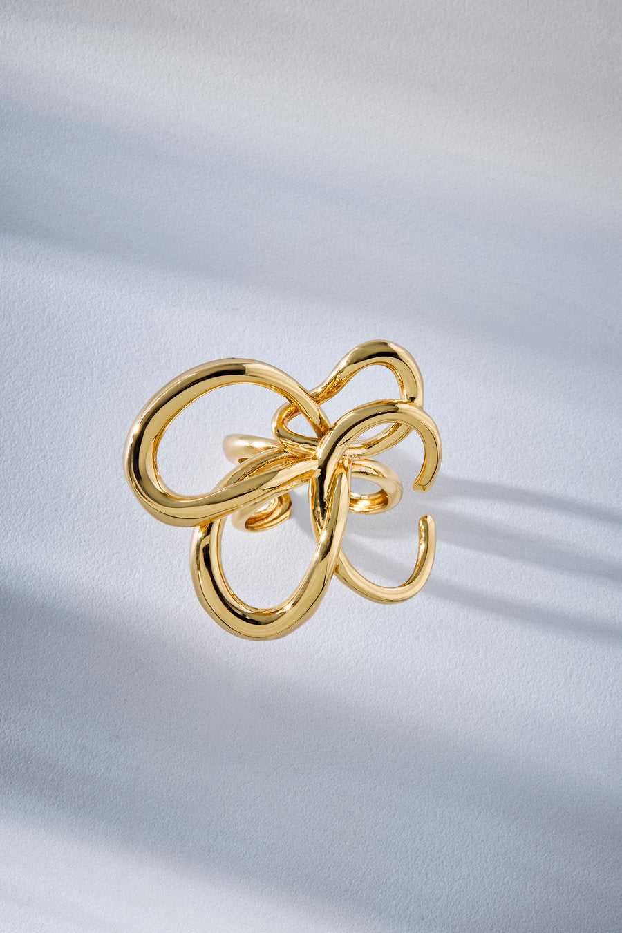 A gold-toned open ring with an abstract, intertwined loop design, photographed on a softly lit white fabric background.