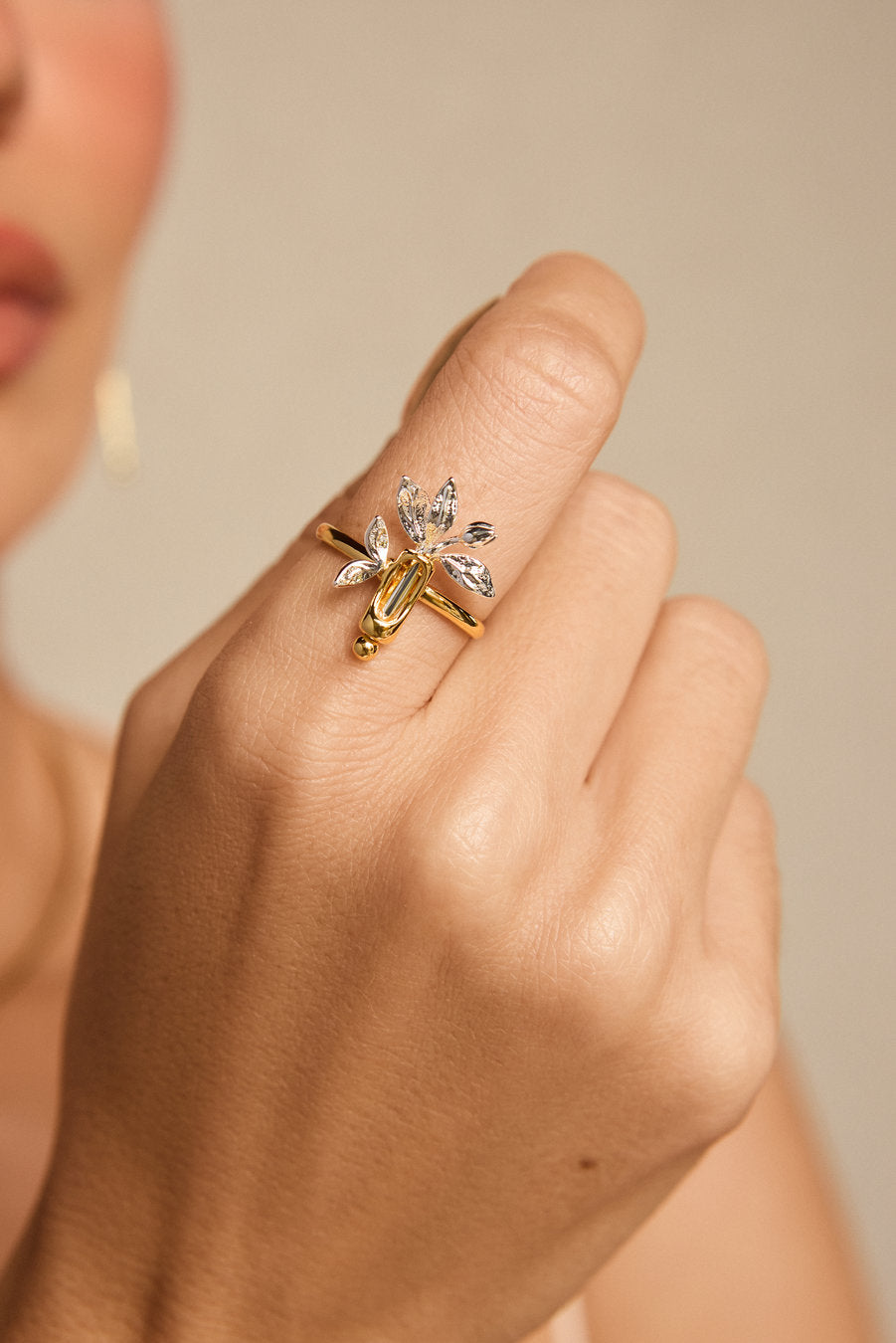 A close-up lifestyle shot of a gold ring with clear crystal leaves, worn on a person's finger against a soft beige background.