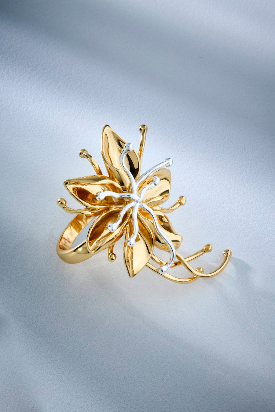 A gold flower-shaped ring with delicate curved wire details and small clear stones, photographed on a soft light gray fabric background.