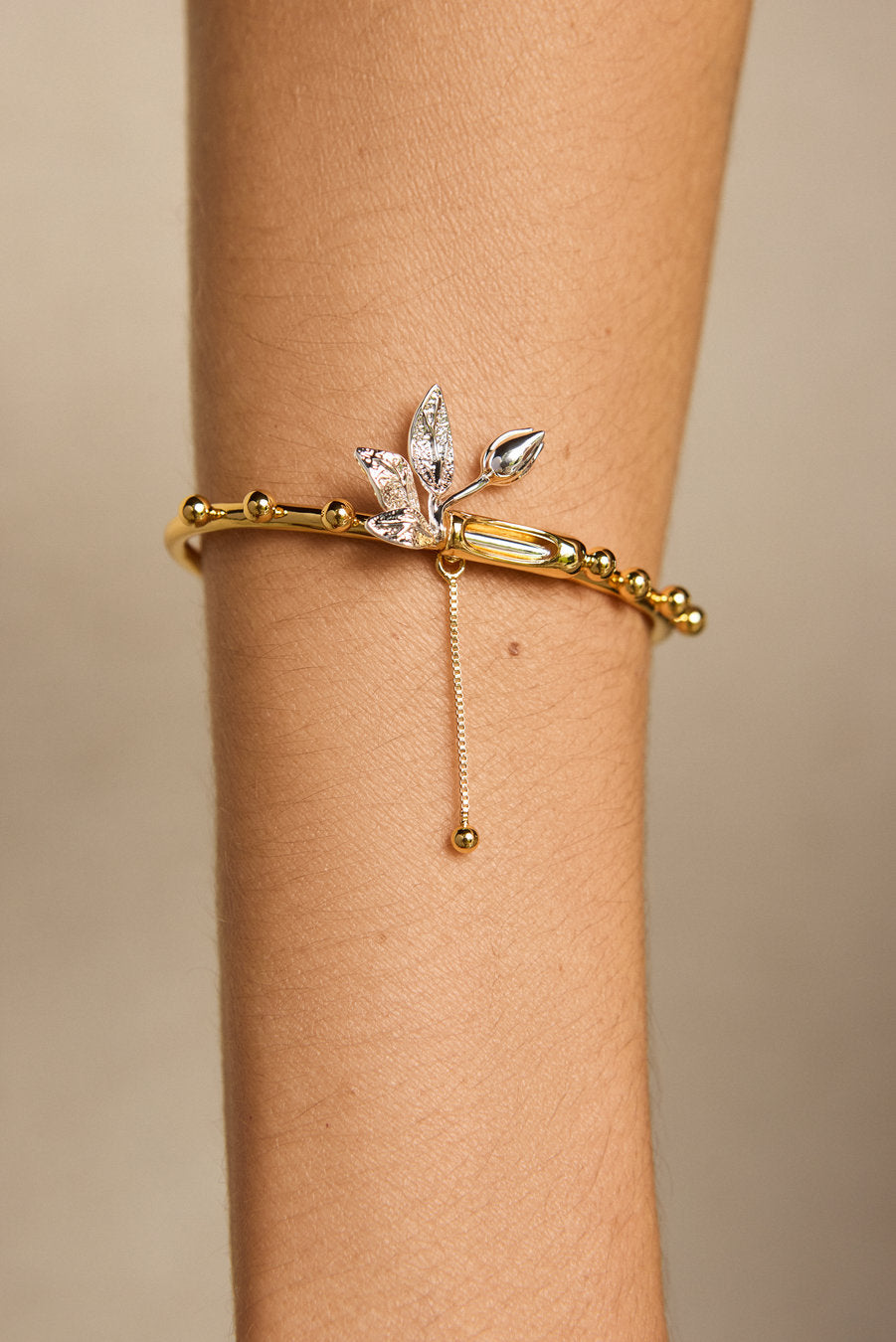 A gold bangle bracelet with a dangling silver-toned leaf charm and clear crystal accents, shown worn on a person's wrist against a soft beige background.