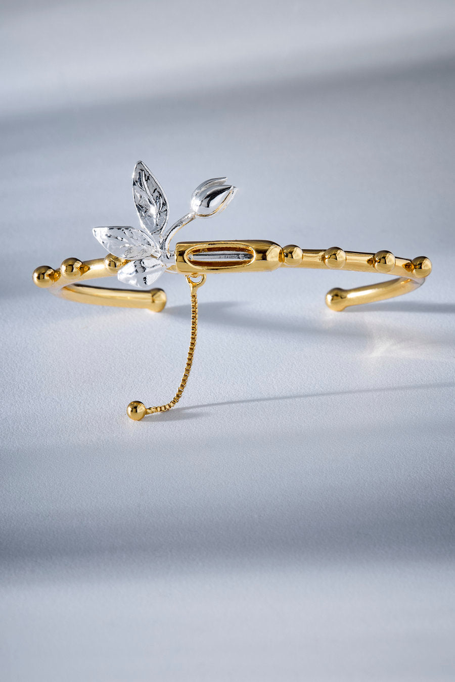 A gold-toned open cuff bracelet featuring a silver-toned leaf and flower motif with clear rhinestone accents, displayed on a light gray surface.