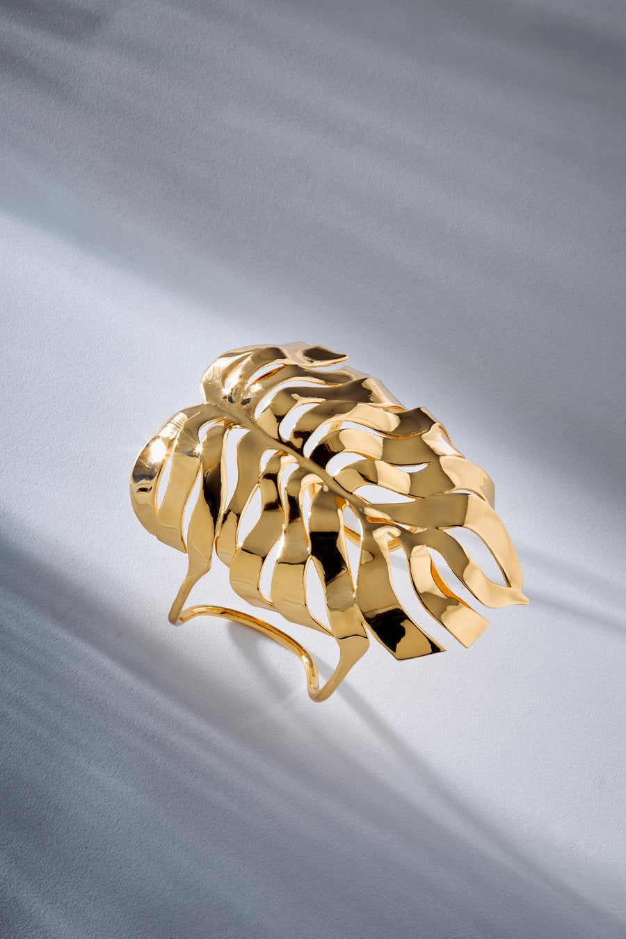 A polished gold ring sculpted in the shape of a monstera leaf, displayed on a draped light gray satin fabric.