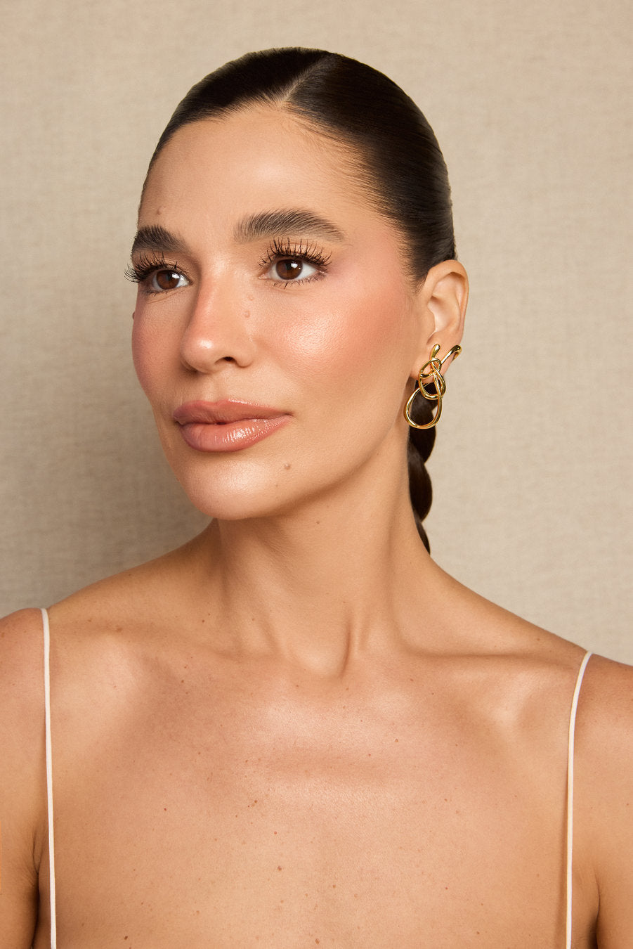 A close-up portrait of a woman with dark hair pulled back, wearing a pair of gold hoop earrings with a textured, layered circular design. She is wearing a white spaghetti-strap top against a neutral beige textured background.