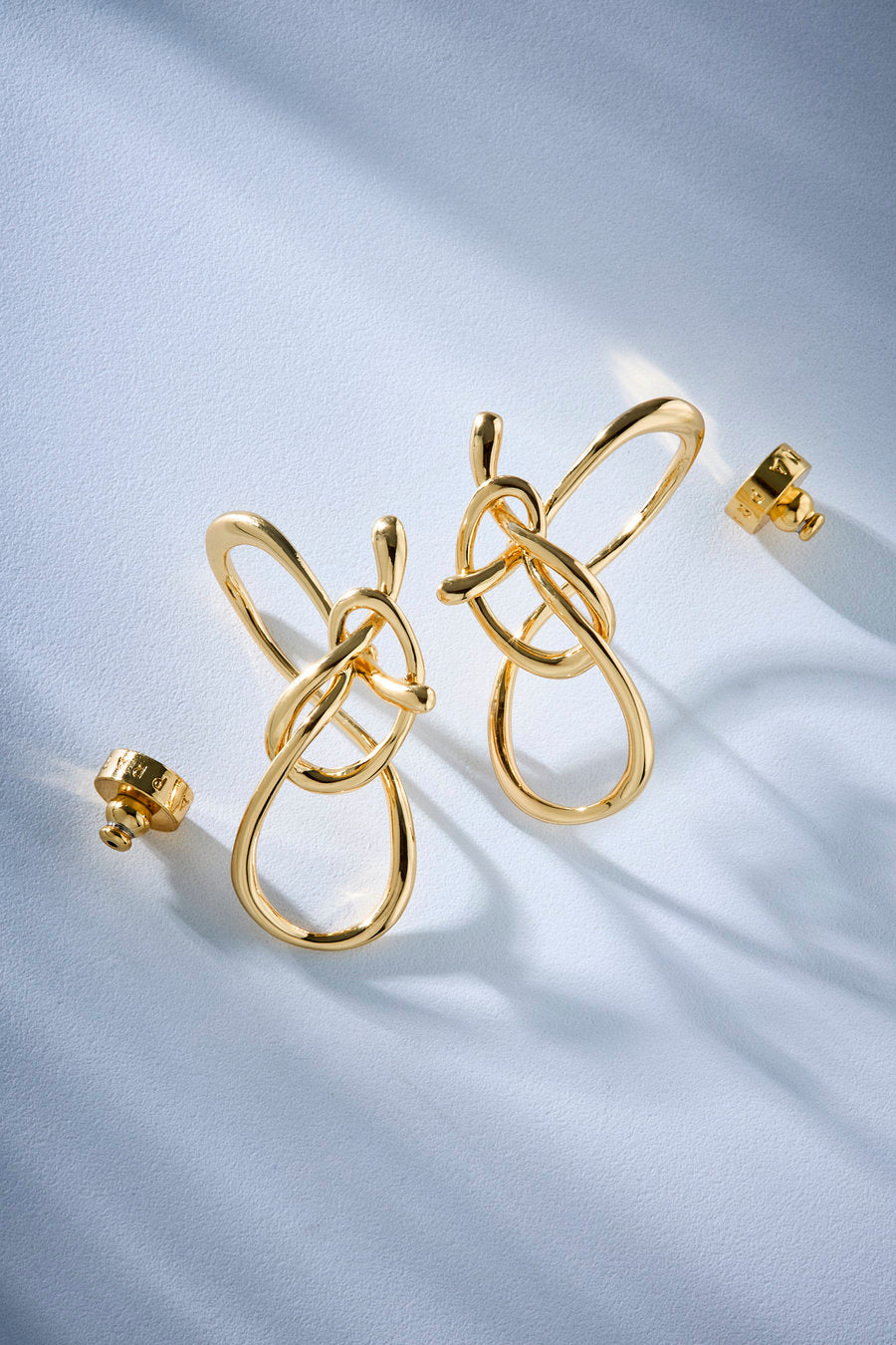 Gold-toned statement earrings with an abstract, intertwined loop design, shown on a soft white satin-like surface with a few matching small pieces nearby.
