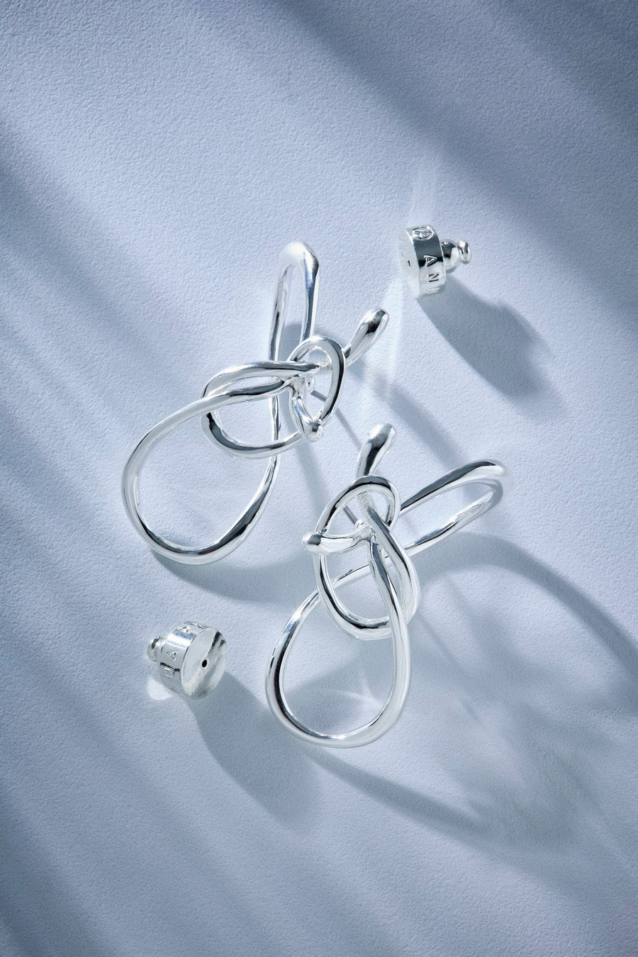 A pair of silver-toned abstract knot earrings with matching stud backs, displayed on a textured light blue surface with soft shadows.