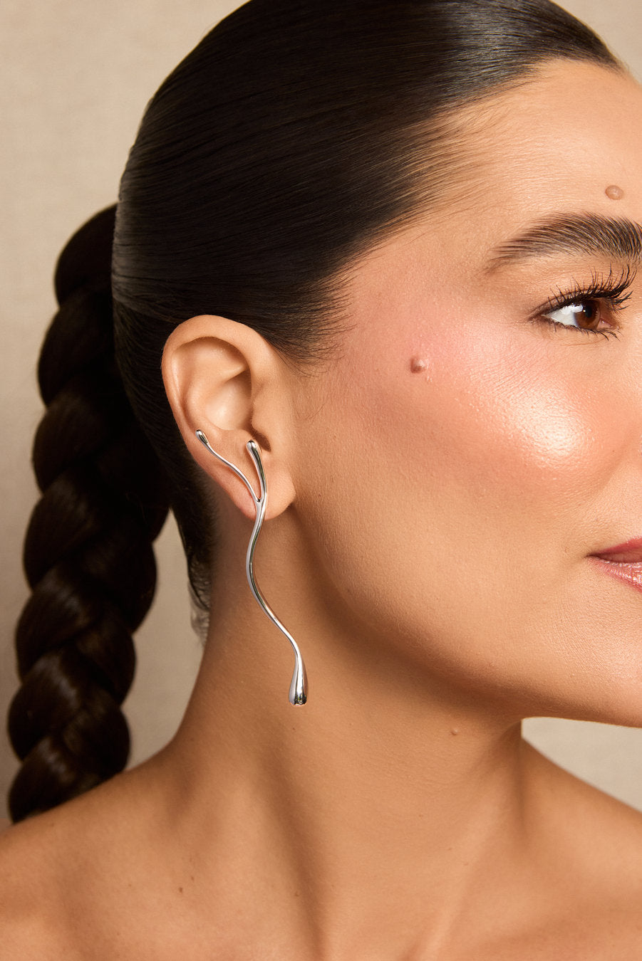 A close-up of a woman with dark hair in a braid wearing a single silver ear cuff earring. The earring has a curved, snake-like form that wraps around the ear and hangs down.