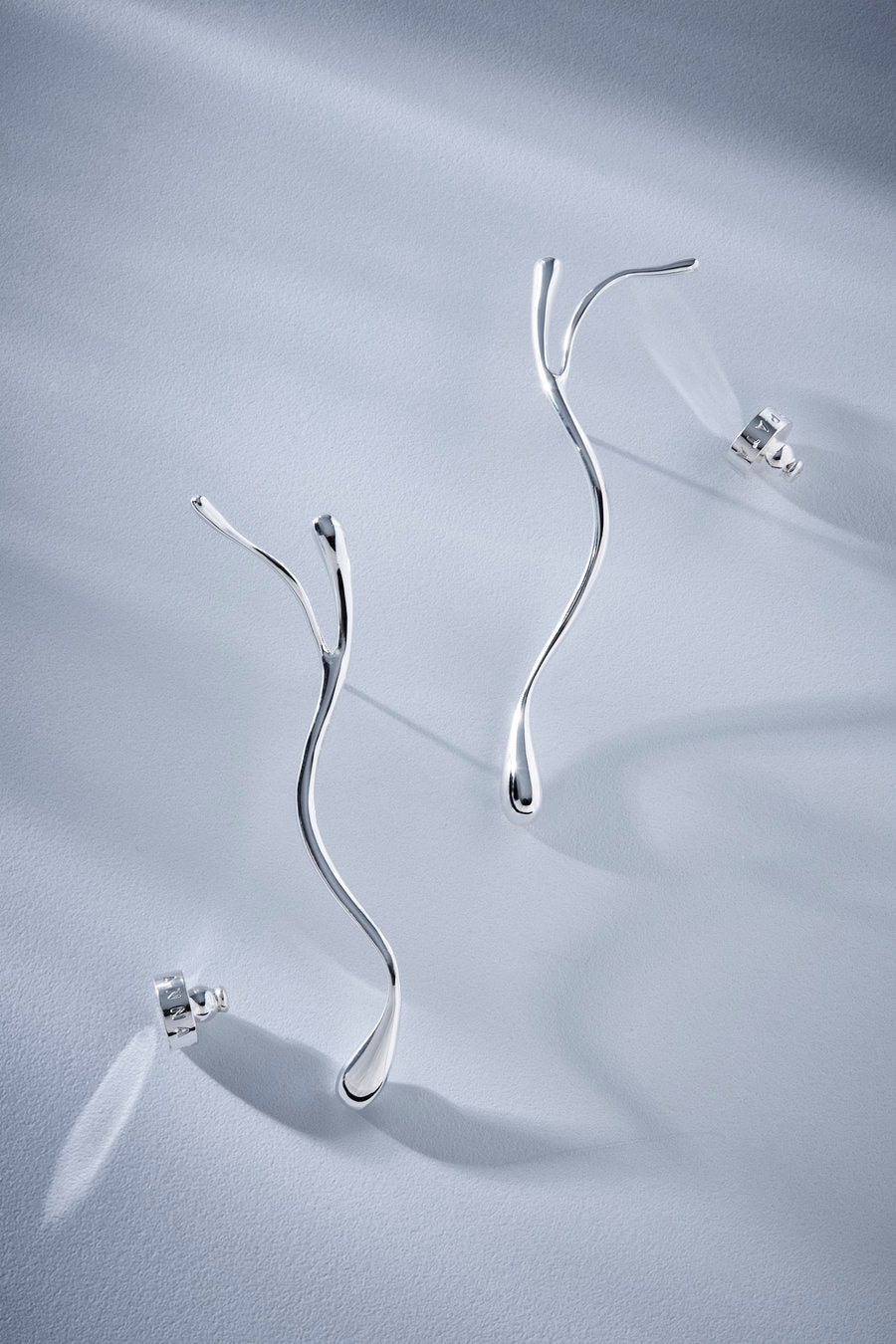 A pair of sculptural, wavy silver earrings with post backs, displayed on a textured light gray surface with soft shadows.