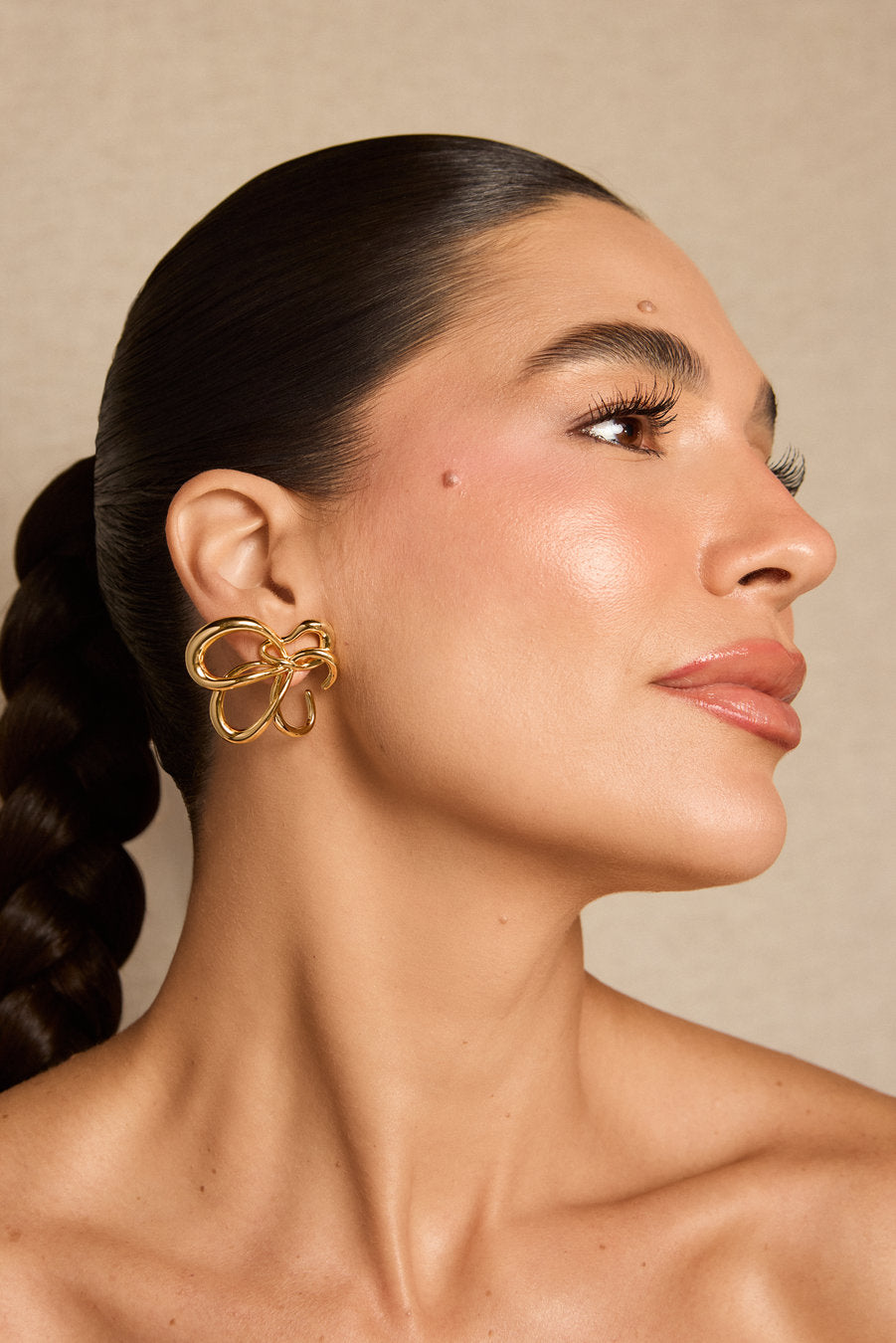 A close-up side profile of a woman with dark hair in a low braid wearing a large, gold bow-shaped hoop earring against a warm beige background.