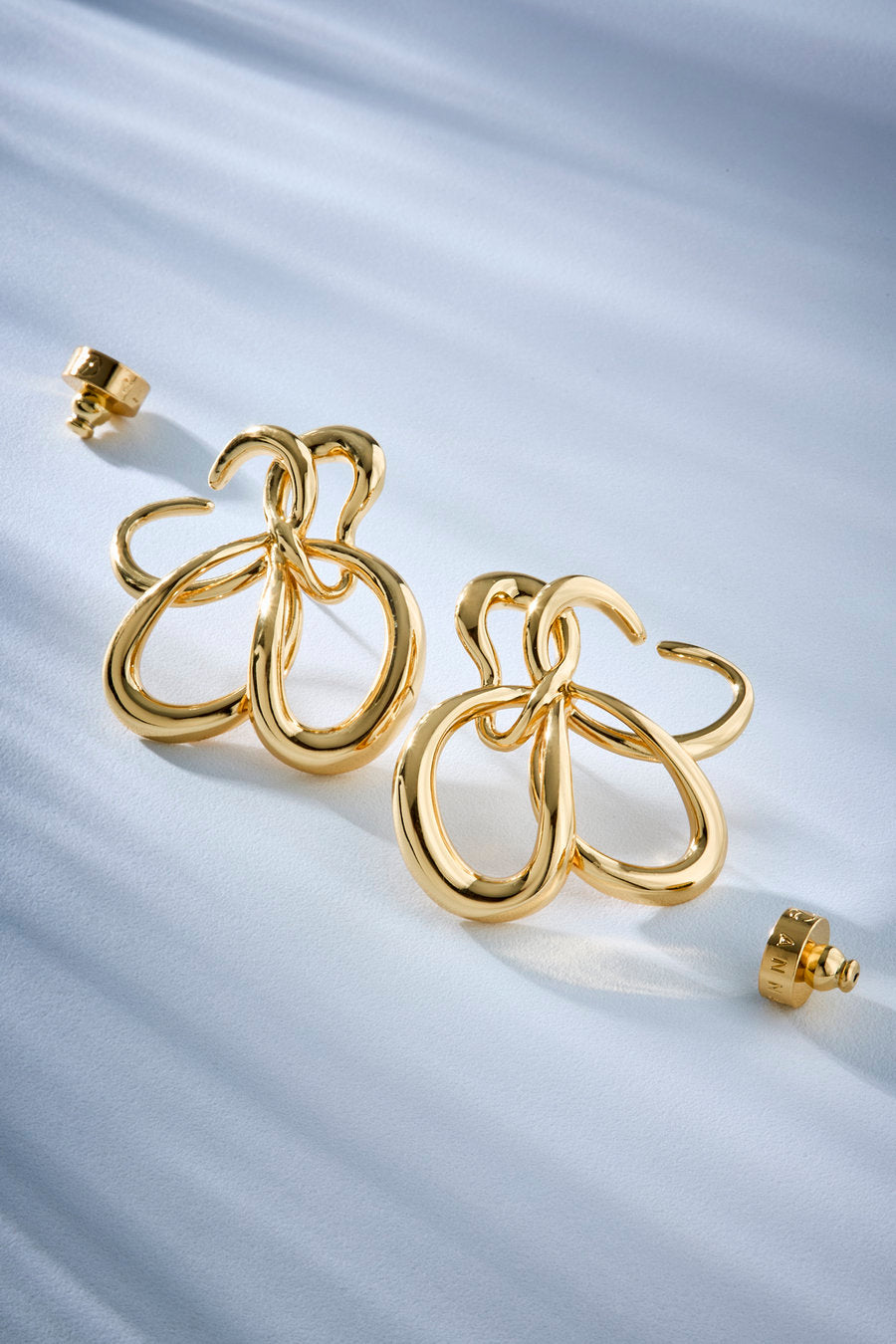 A pair of gold-toned statement earrings with an abstract, openwork looped design, displayed on a softly draped light blue fabric background.