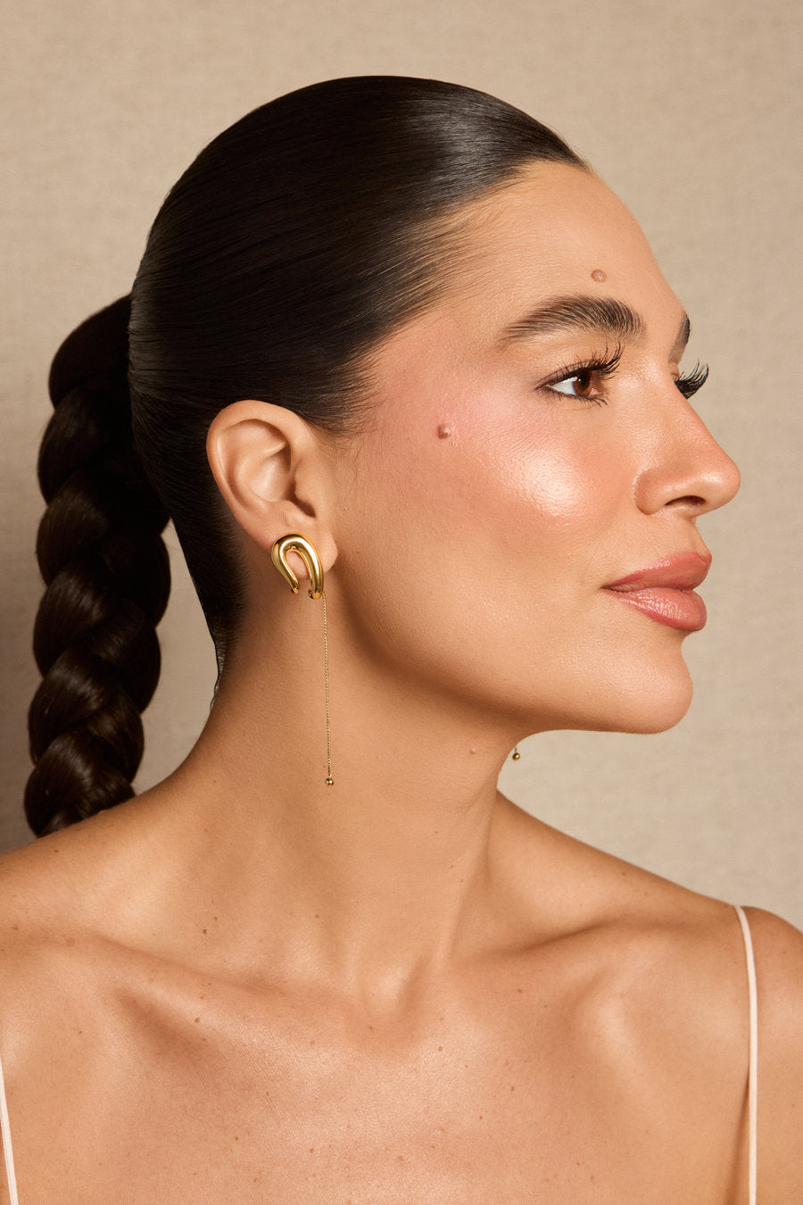 A woman in profile wearing a gold crescent-shaped hoop earring with a long dangling chain, shown against a warm beige background.